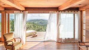 A wooden room with a bed, a chair, and a view of a mountainous landscape through large windows. - Castello Di Vicarello (Cinigiano)
