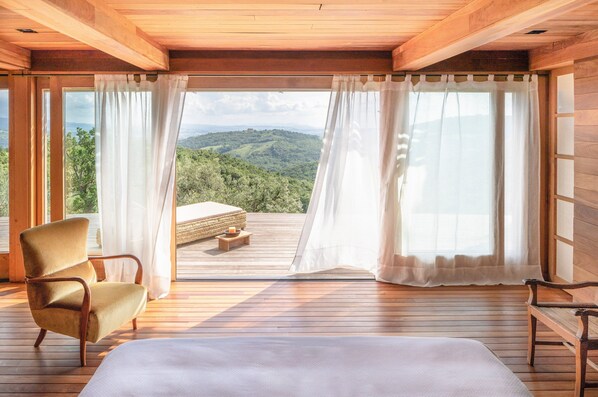 A wooden room with a bed, a chair, and a view of a mountainous landscape through large windows.