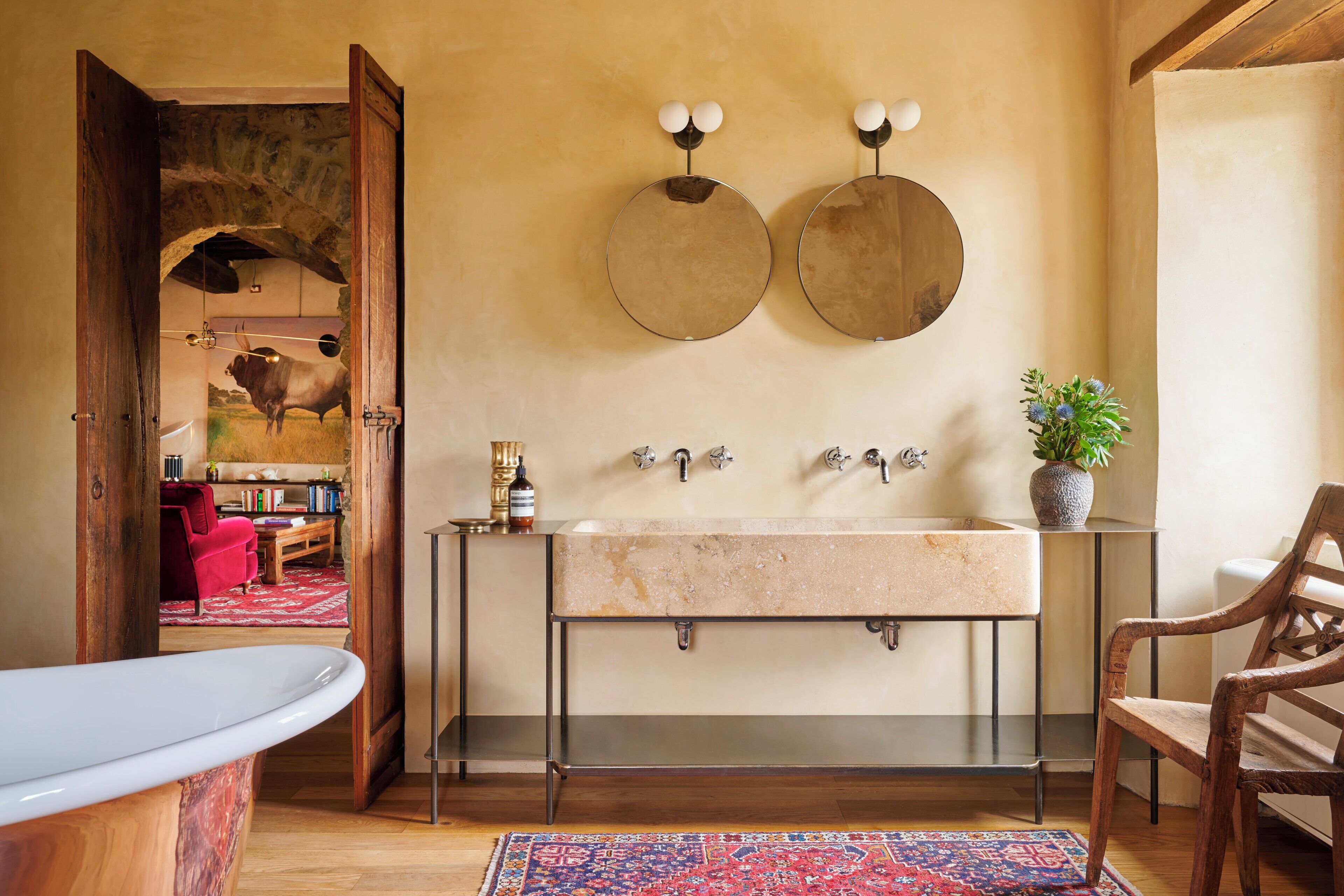 Un baño con un lavamanos grande, dos espejos redondos, una silla de madera y un tapete rojo.