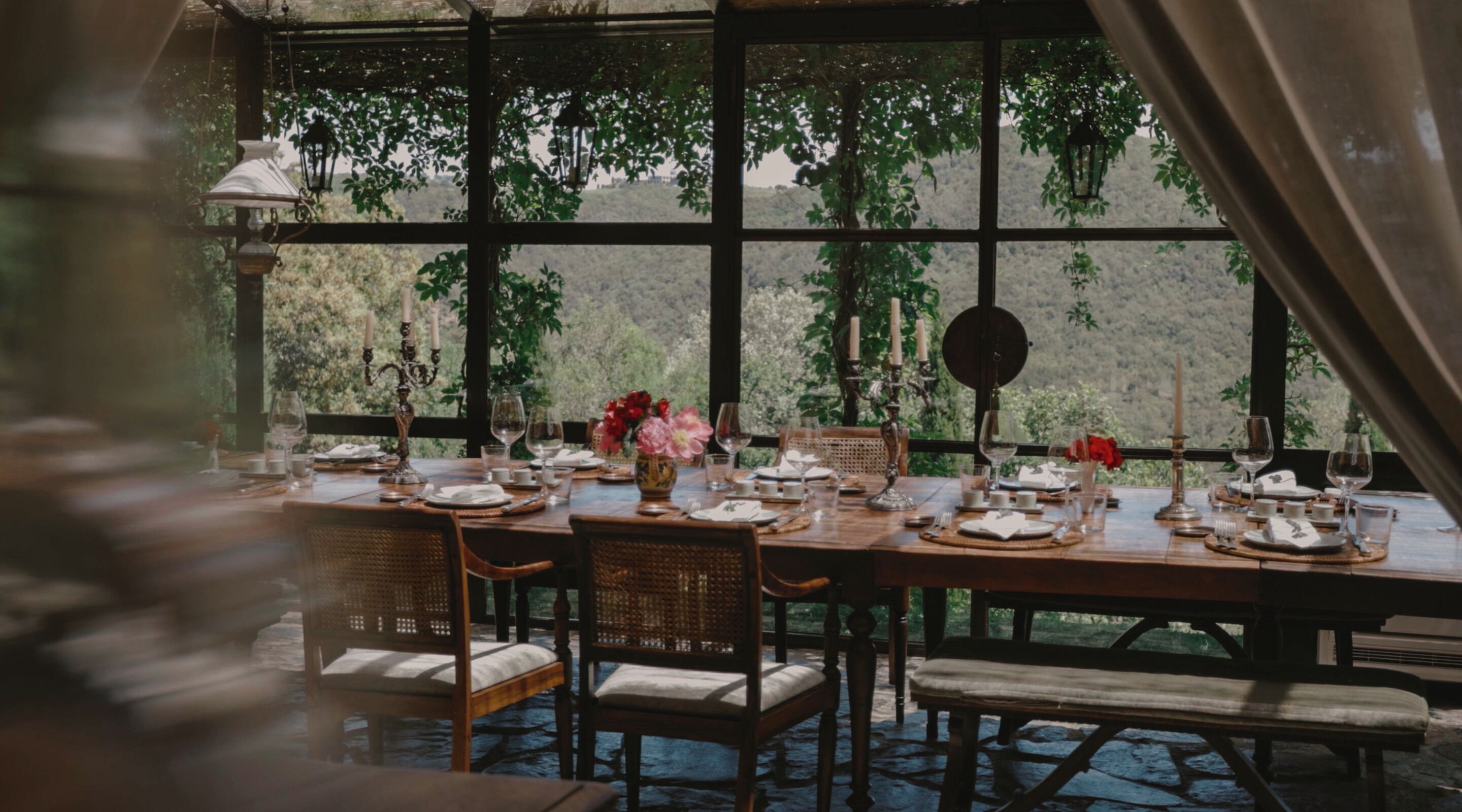 Mesa de comedor lista para seis personas, con velas, flores y vistas a árboles a través de amplias ventanas.