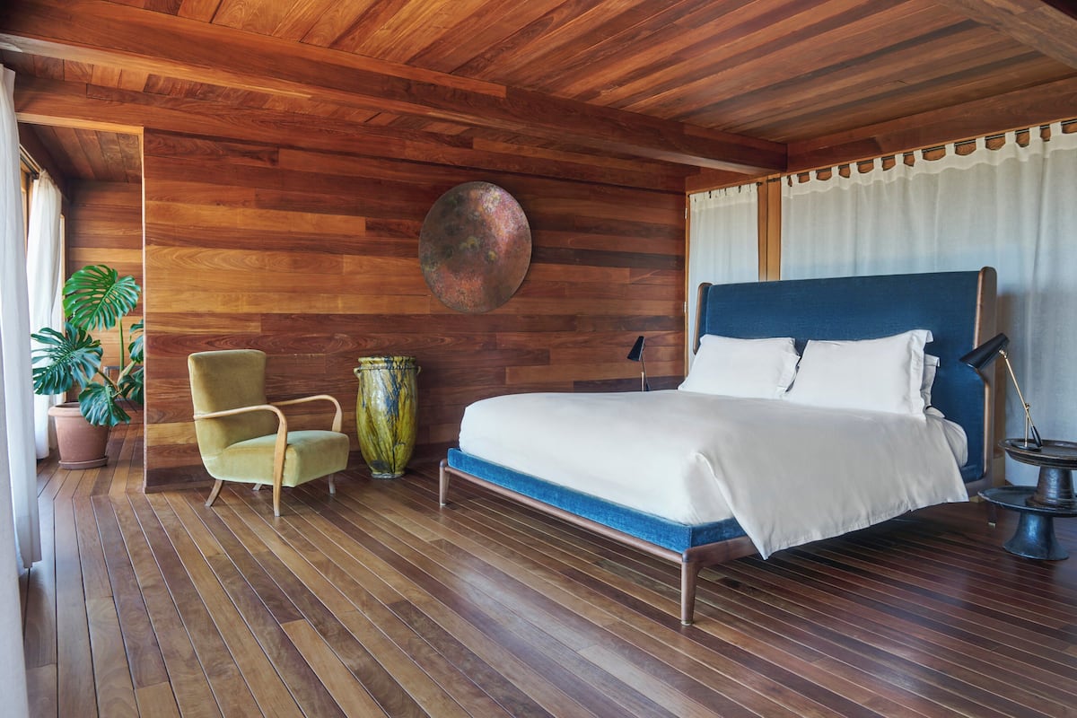 Un dormitorio con piso de madera, una cama con cabecera azul, un sillón verde y un jarrón grande.