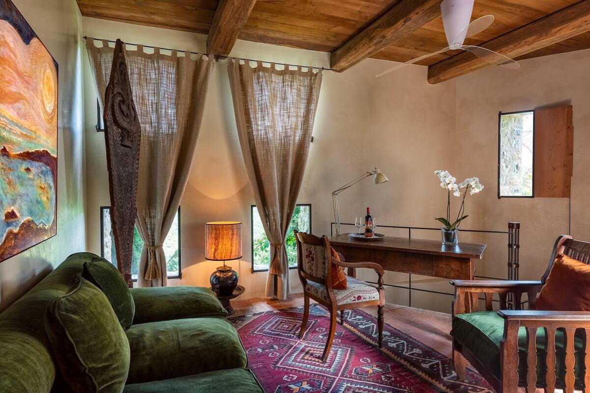 Una habitación acogedora con un sofá verde, un escritorio de madera y una silla. Hay una lámpara, una planta y un cuadro en la pared.