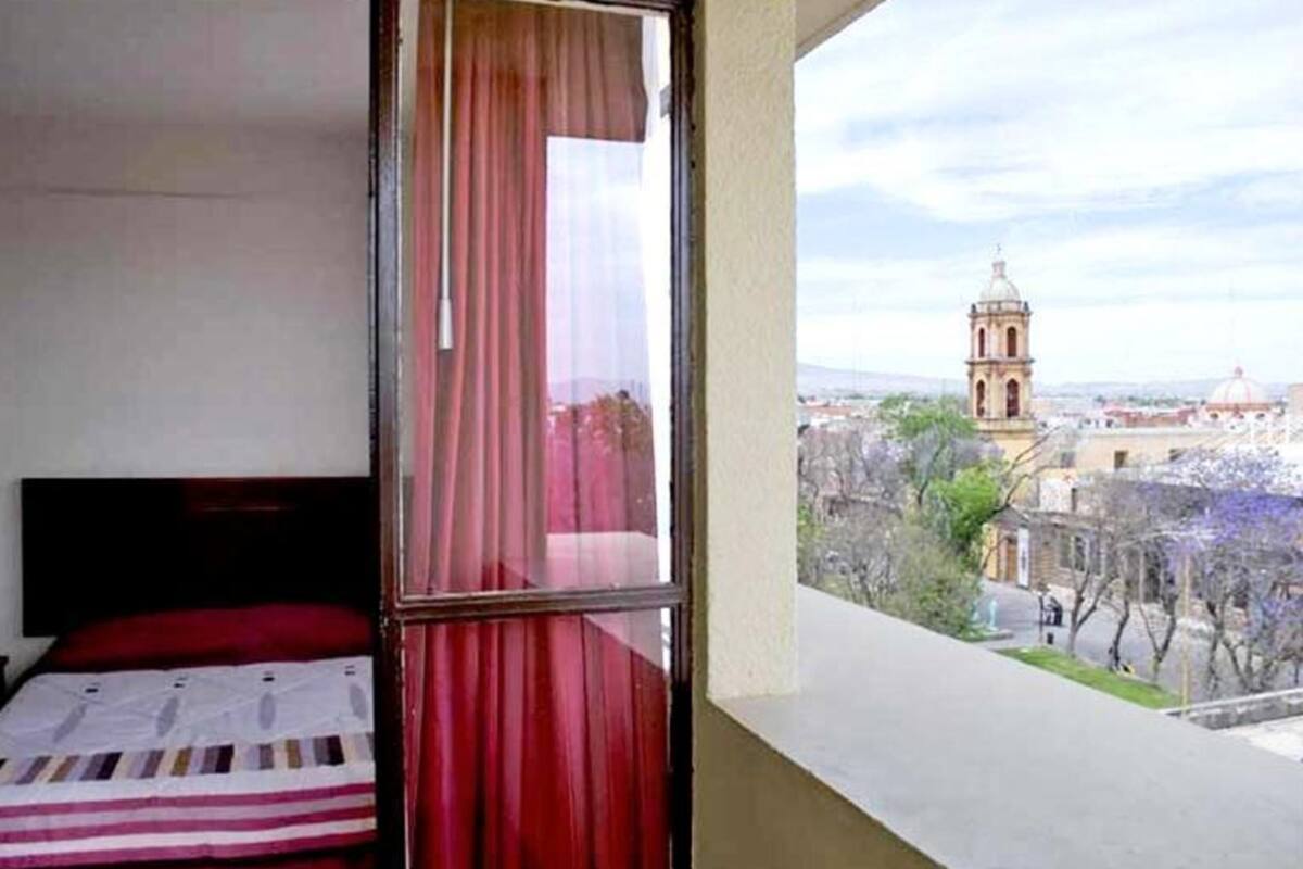 A room with a bed, a window with red curtains, and a view of a cityscape with a prominent building.