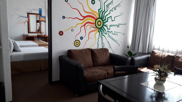 A hotel room with a bed, a brown leather sofa, a dining table with a vase of flowers, and a wall with a colorful abstract design.
