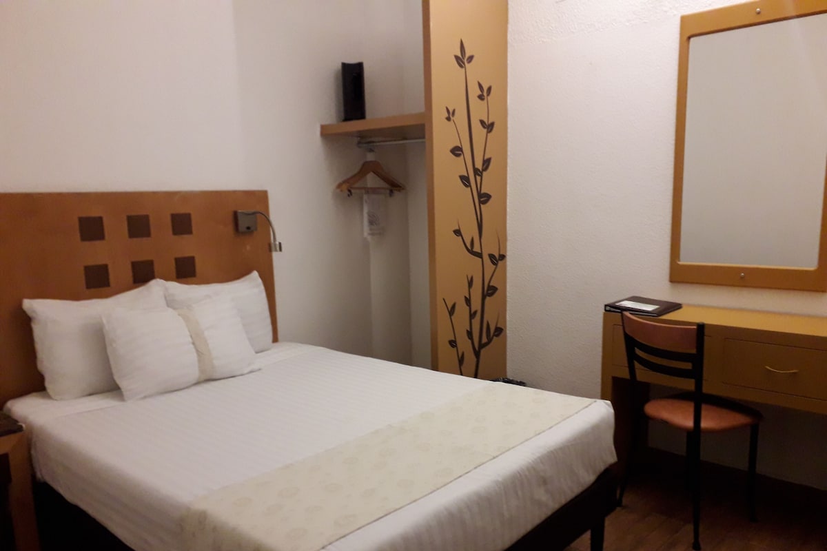 Een net opgemaakt bed met hoofdbord, een bureau met stoel, en een spiegel aan de muur.