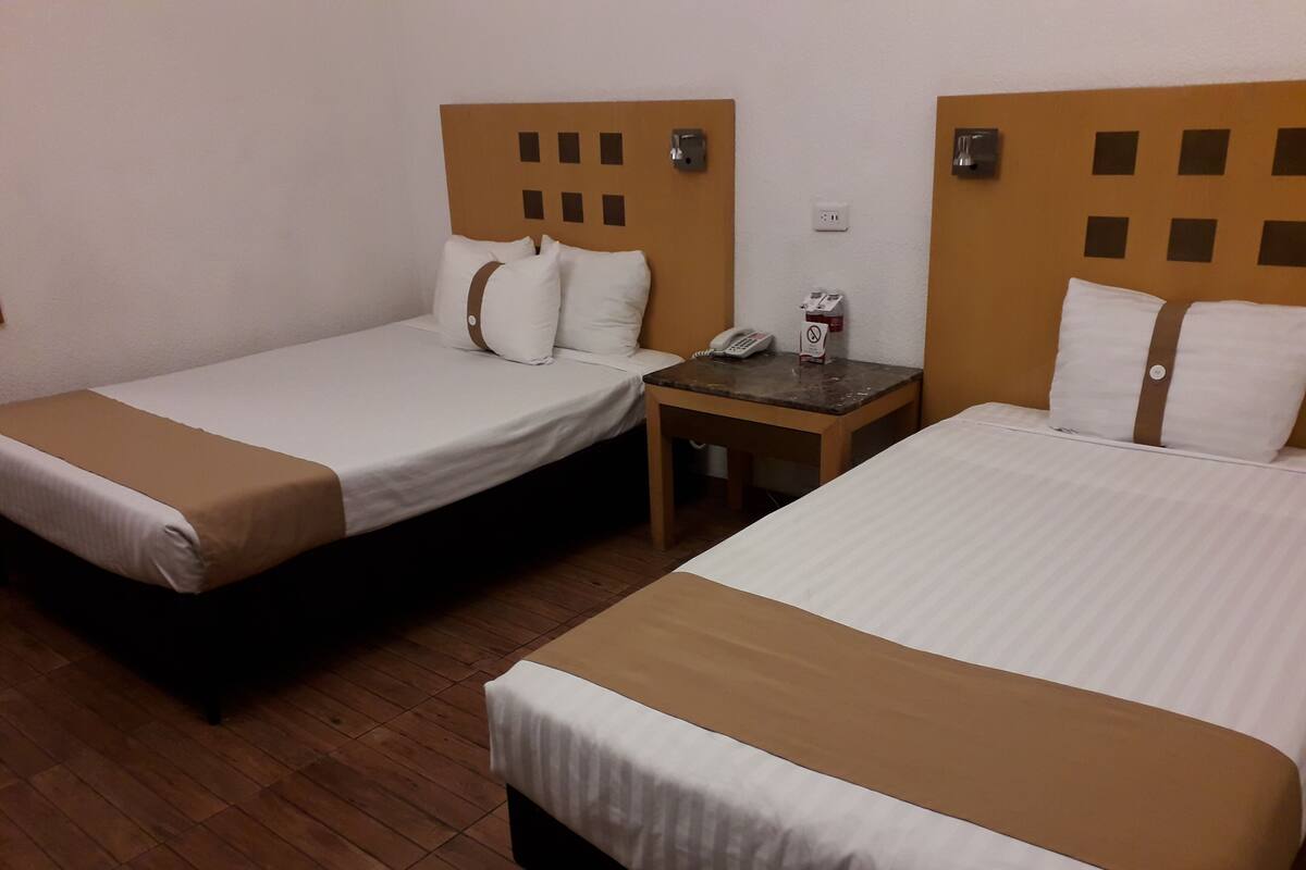 A hotel room with two single beds, each with a headboard featuring a grid pattern. There is a small table with a telephone and a 'No Smoking' sign between the beds.