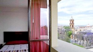A room with a bed, a window with red curtains, and a view of a cityscape including a church tower.