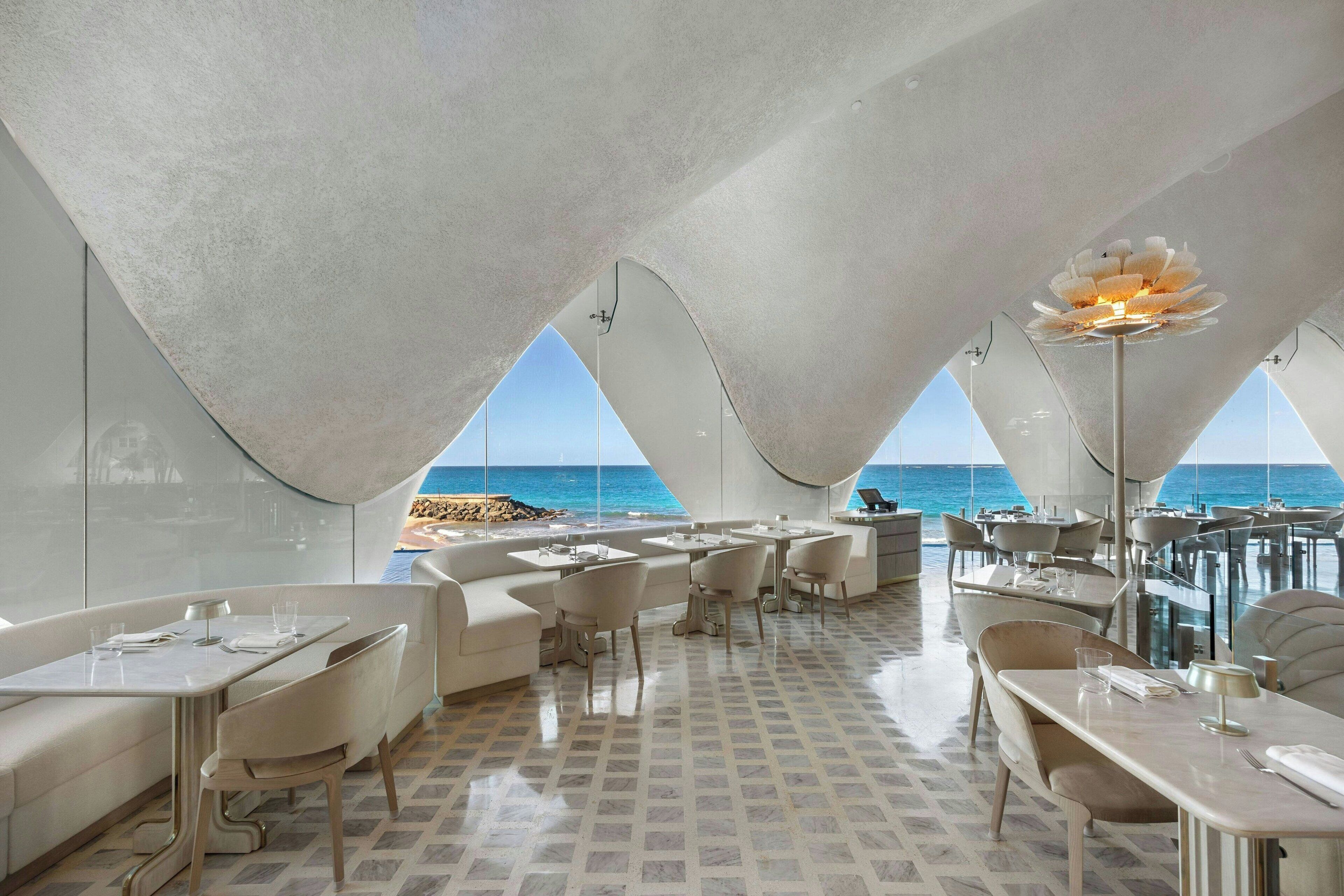 A modern restaurant with a view of the sea, featuring a unique ceiling design and a central flower-shaped light fixture.