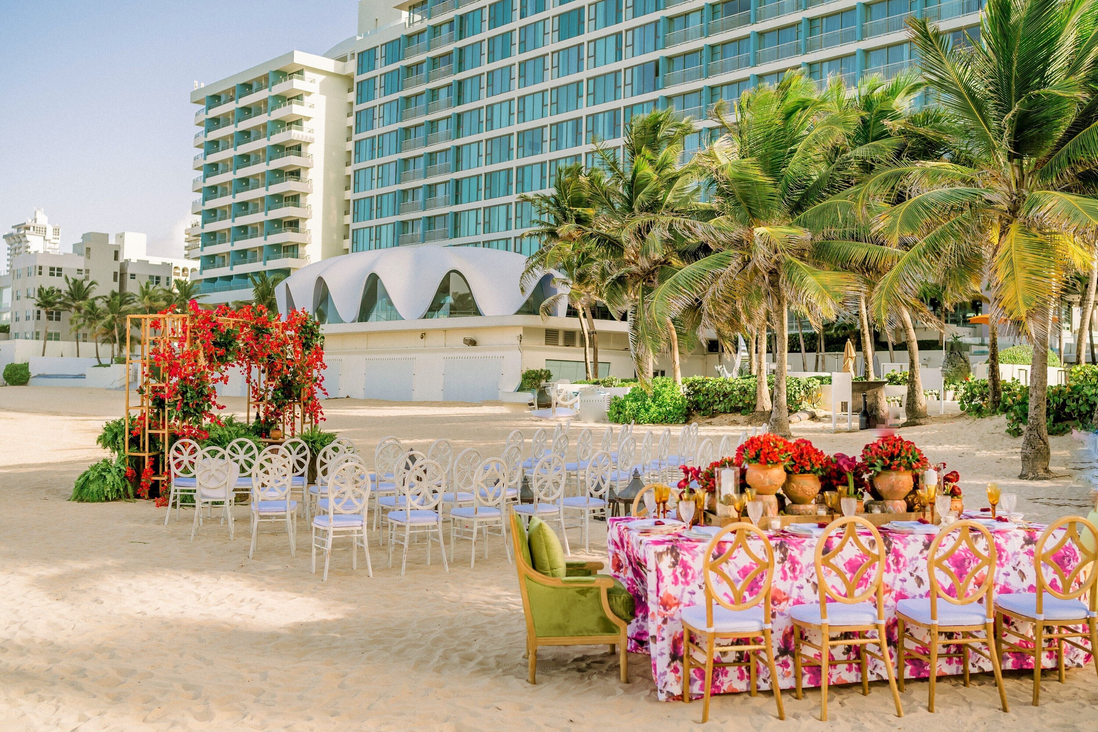 A beachside outdoor dining area with white chairs and tables, palm trees, and a modern building in the background.