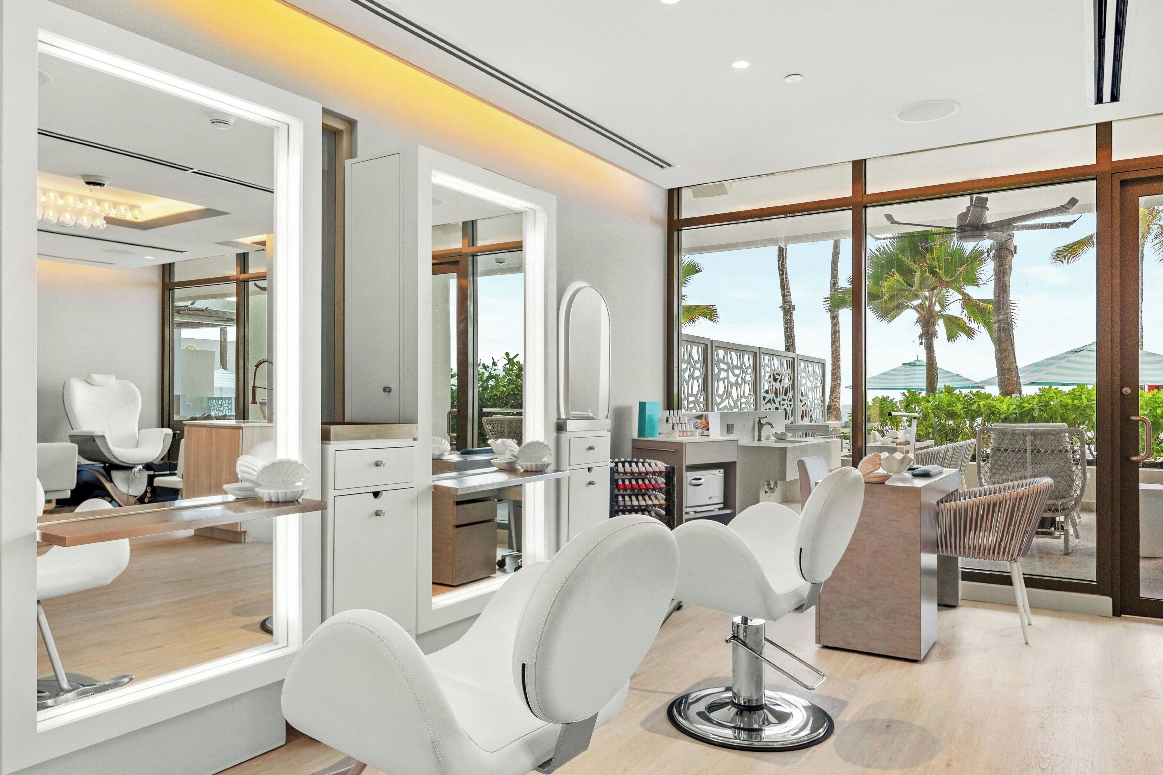 Un salón moderno con sillas blancas, un espejo grande y vistas a palmeras a través de amplios ventanales.