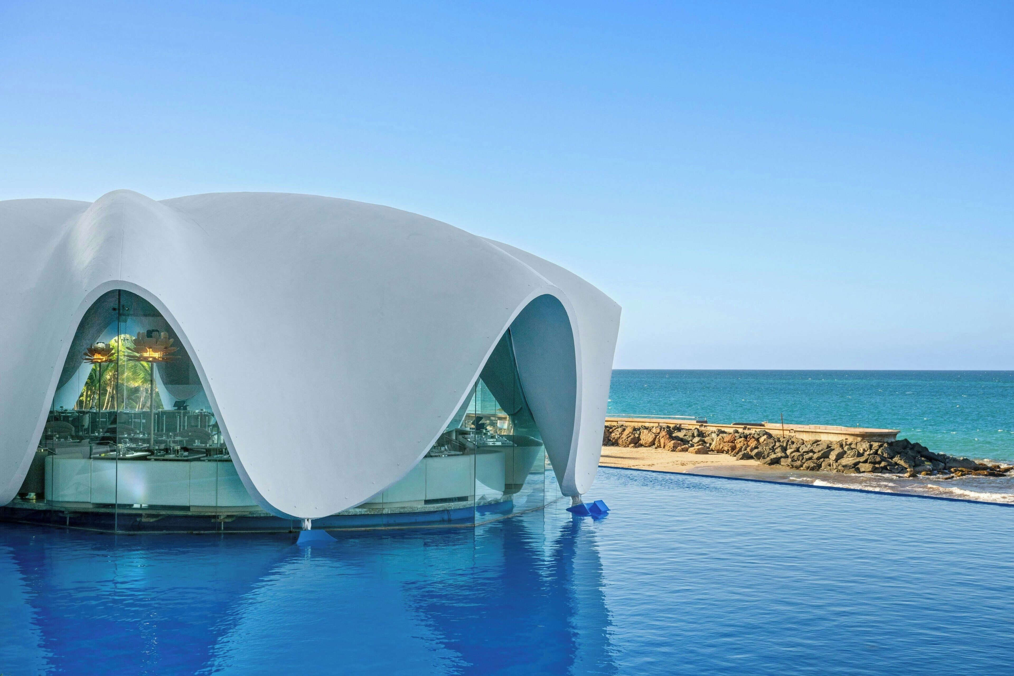Un edificio moderno con un diseño de techo curvo único, ubicado junto a una piscina con vista al océano.