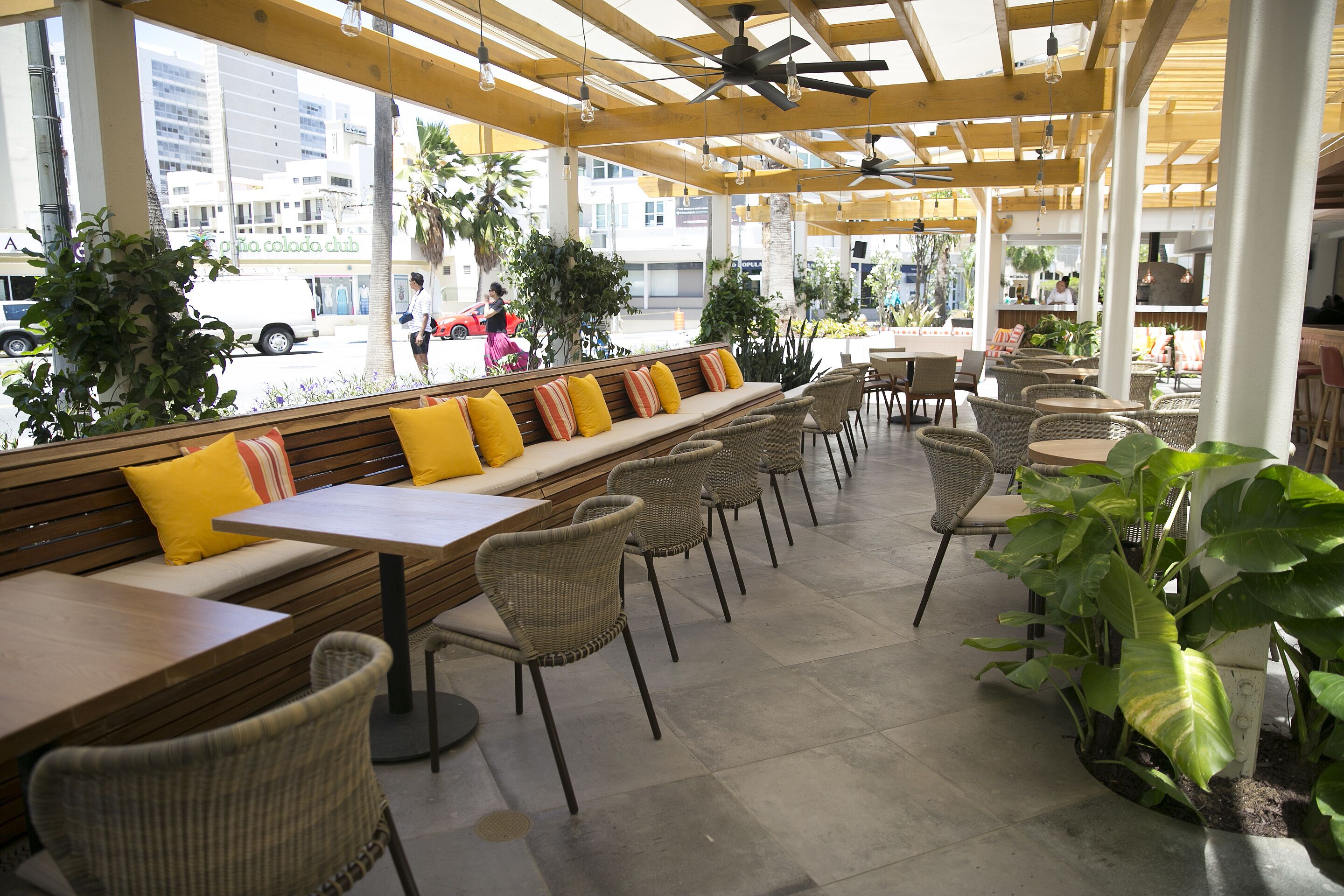 An outdoor cafe with wooden benches, wicker chairs, and large windows overlooking a street.