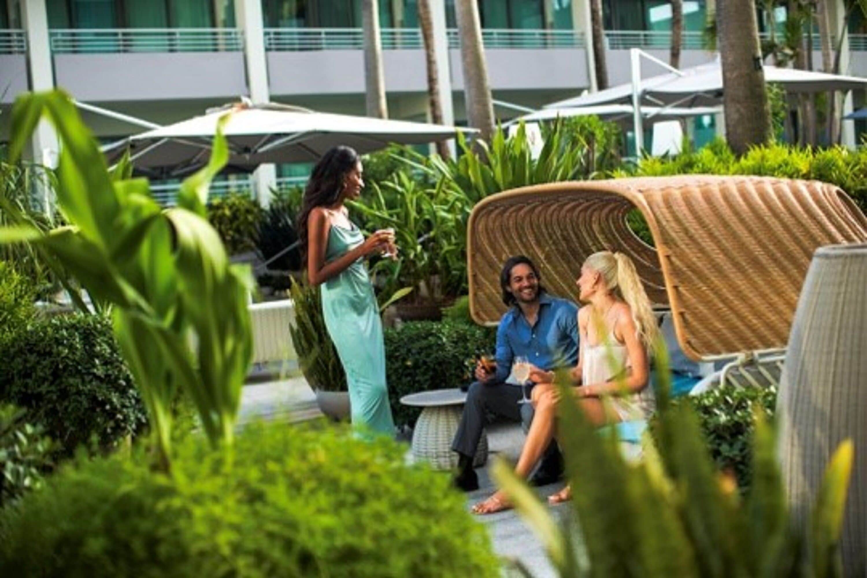 A group of people sitting on outdoor furniture in a lush garden area with a modern building in the background.