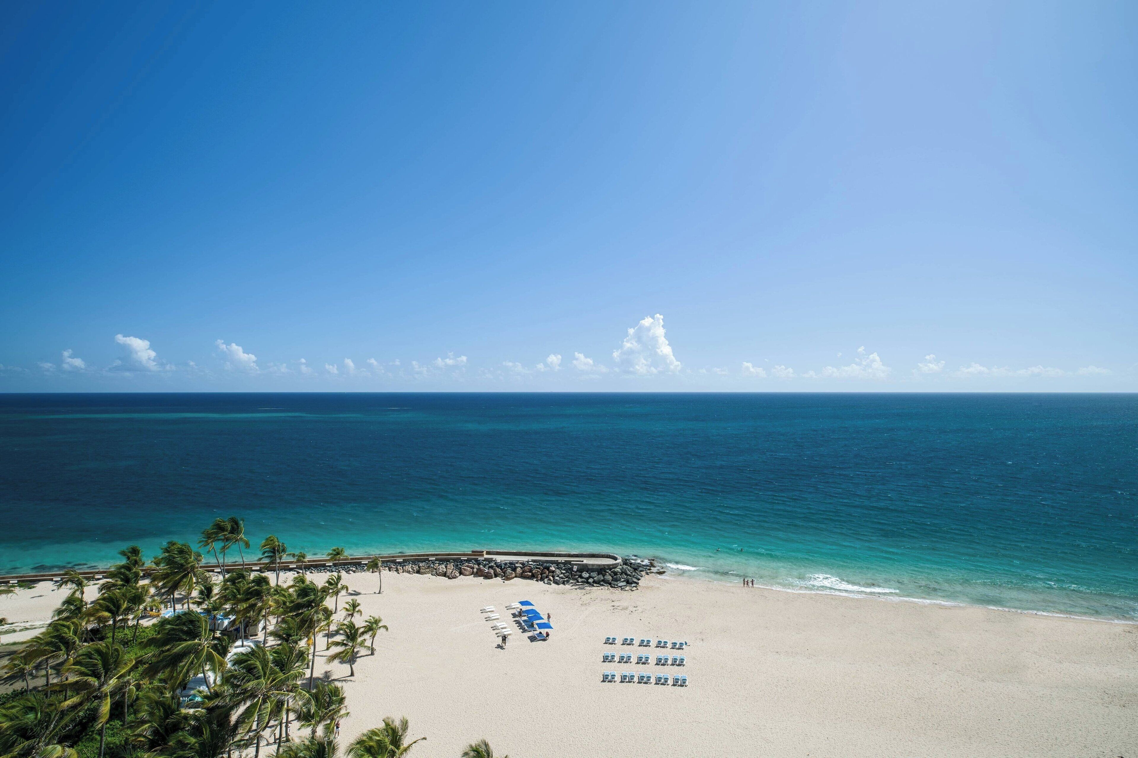 A beach with a clear sky, turquoise water, and palm trees. There are lounge chairs and umbrellas on the sand.