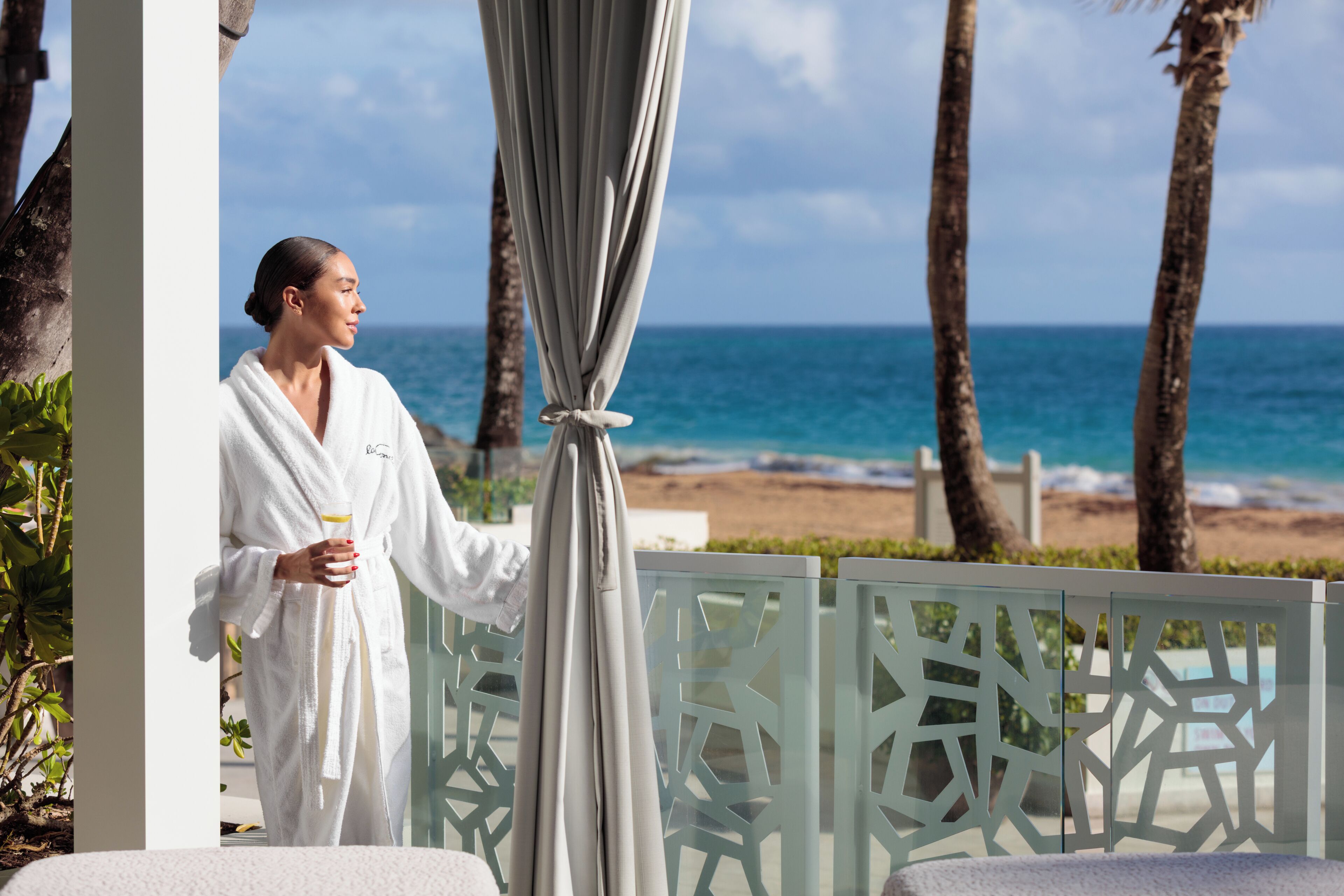 A person in a white robe standing by a glass partition with a view of the beach and ocean.