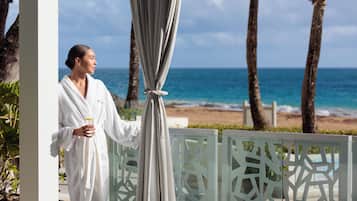 A person in a white robe standing by a glass partition with a view of the beach and ocean.