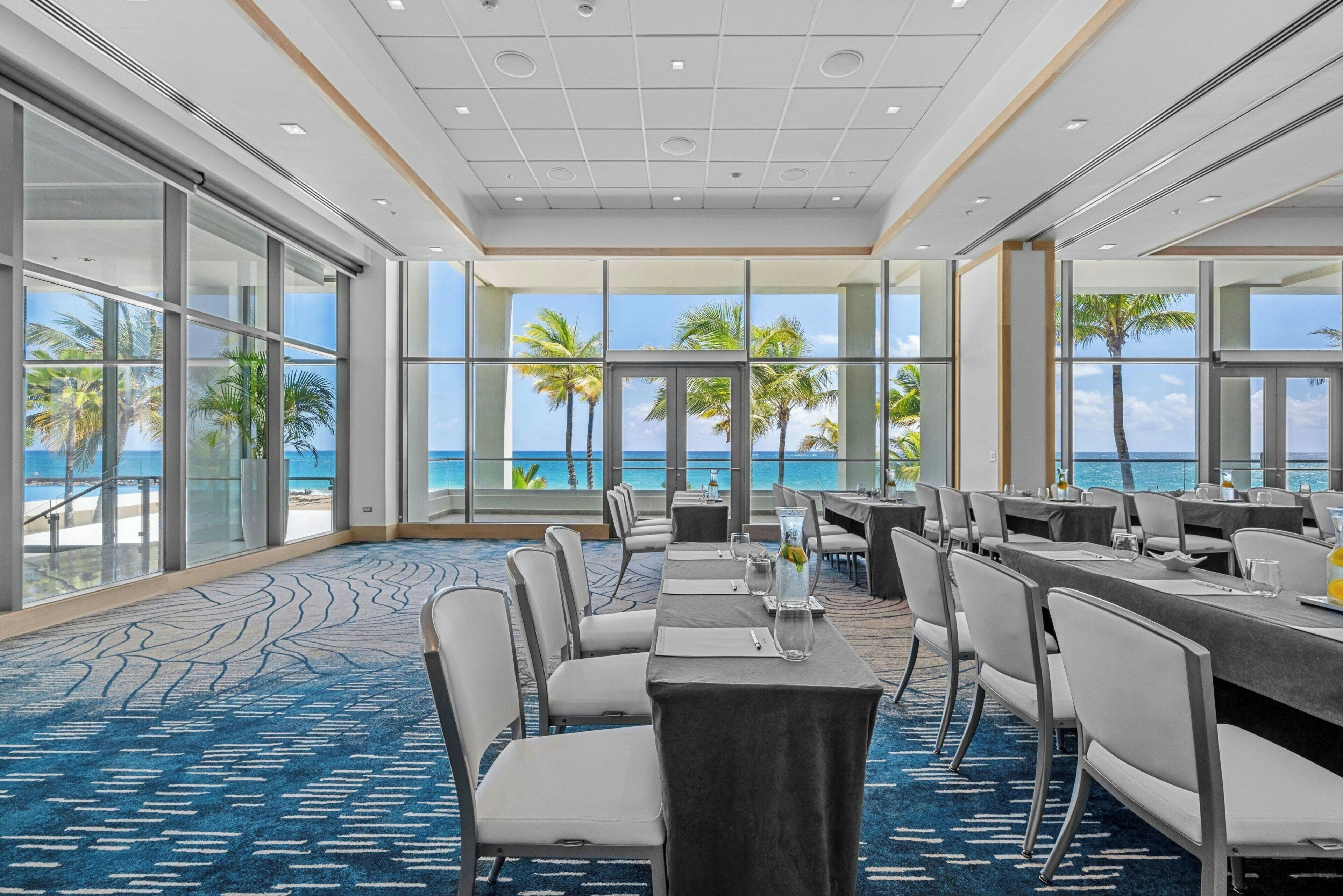 A hotel room with a large window, a view of the ocean, and a blue carpet.