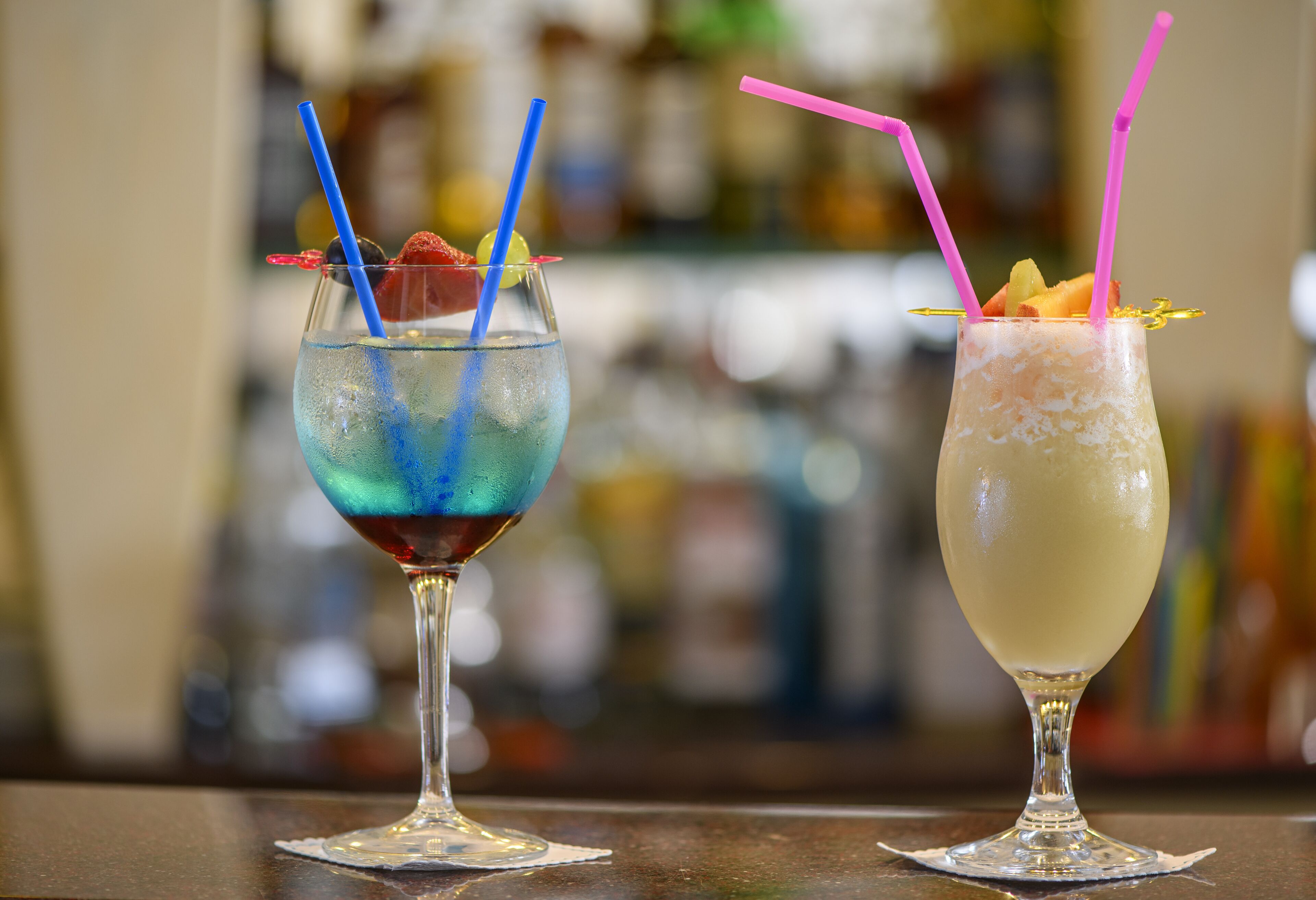 Two cocktails with straws and garnishes on a bar counter.