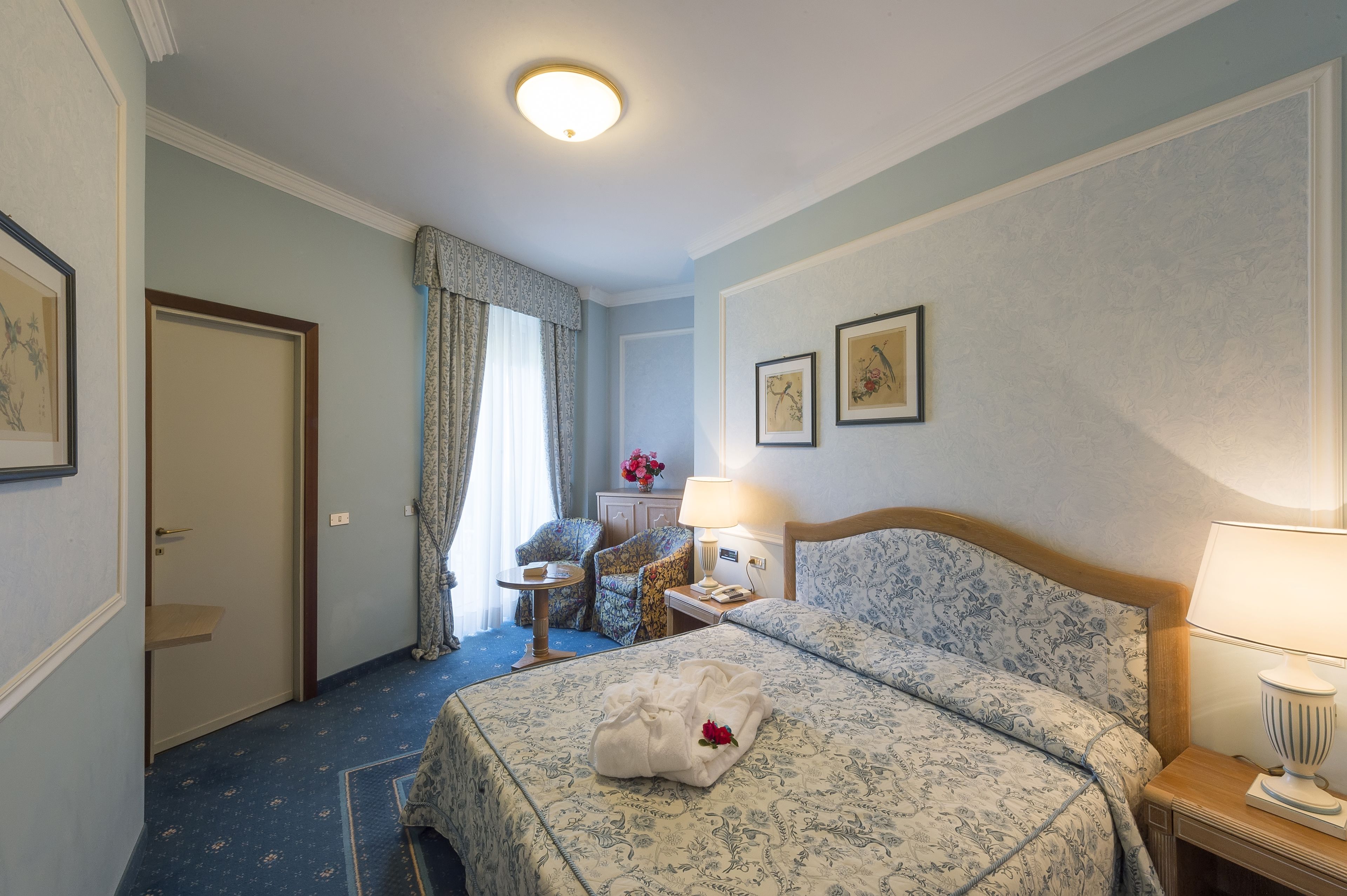 A hotel room with a neatly made bed, a bedside table with a lamp, a chair, a small table with a vase of flowers, and a door leading to another room.