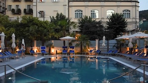 Una piscina con sillas de descanso y sombrillas, rodeada por un edificio con balcones y una cúpula.
