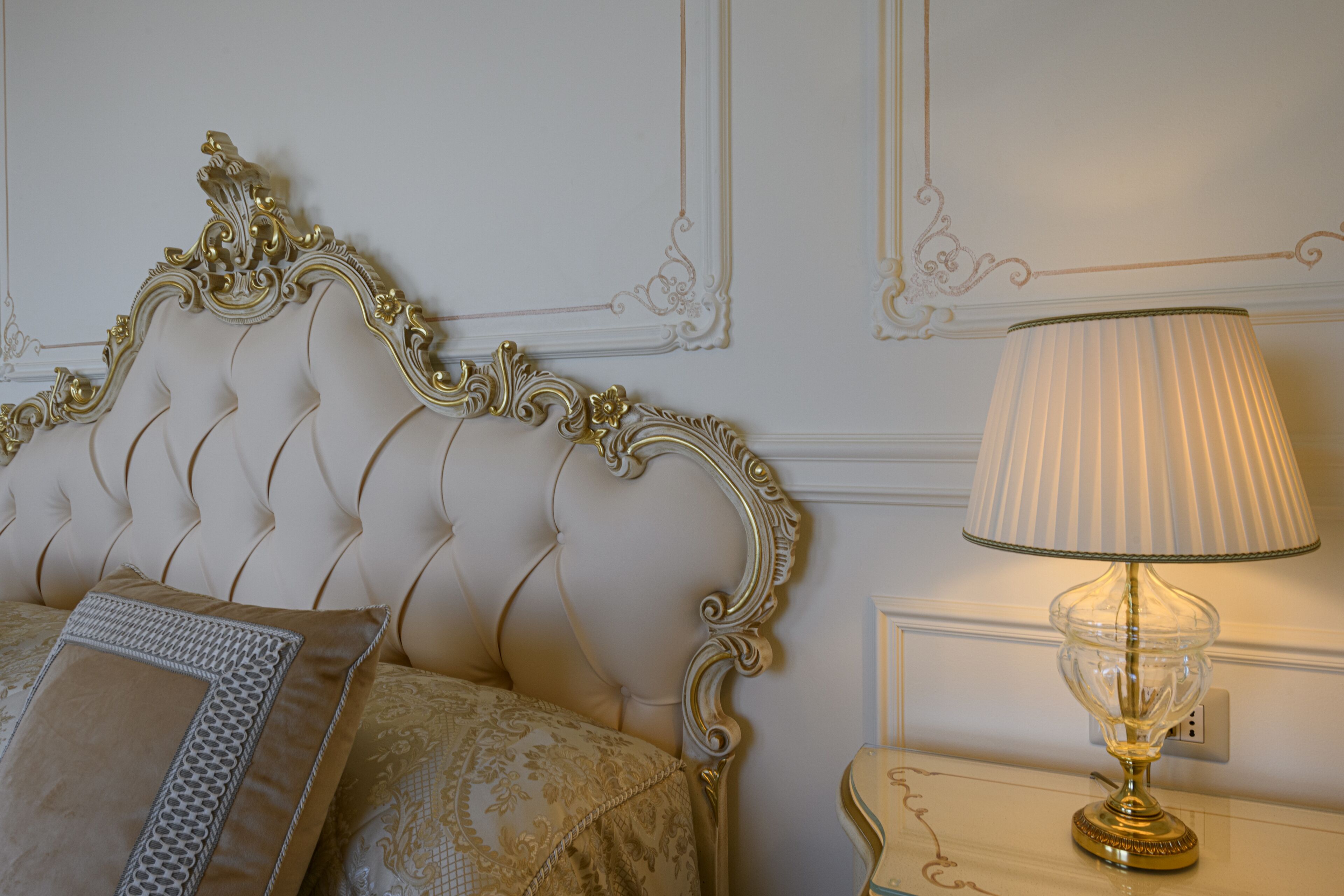 A bed with an ornate headboard, a bedside table with a lamp, and a wall with decorative molding.
