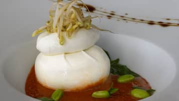 A white plate with a white spherical food item, garnished with green leaves and sprouts, and a red sauce.