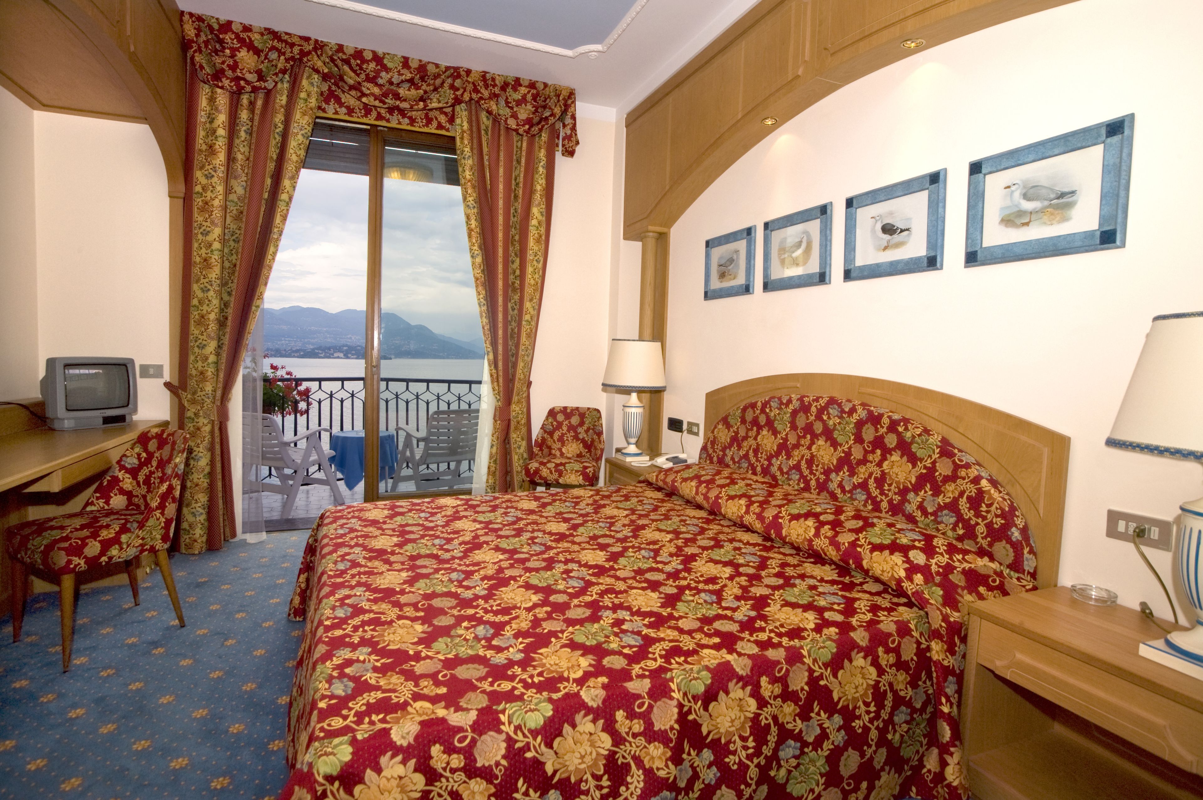 Habitación de hotel con una cama grande, un escritorio con televisor, una silla, un balcón con vistas a las montañas y cuadros en la pared.