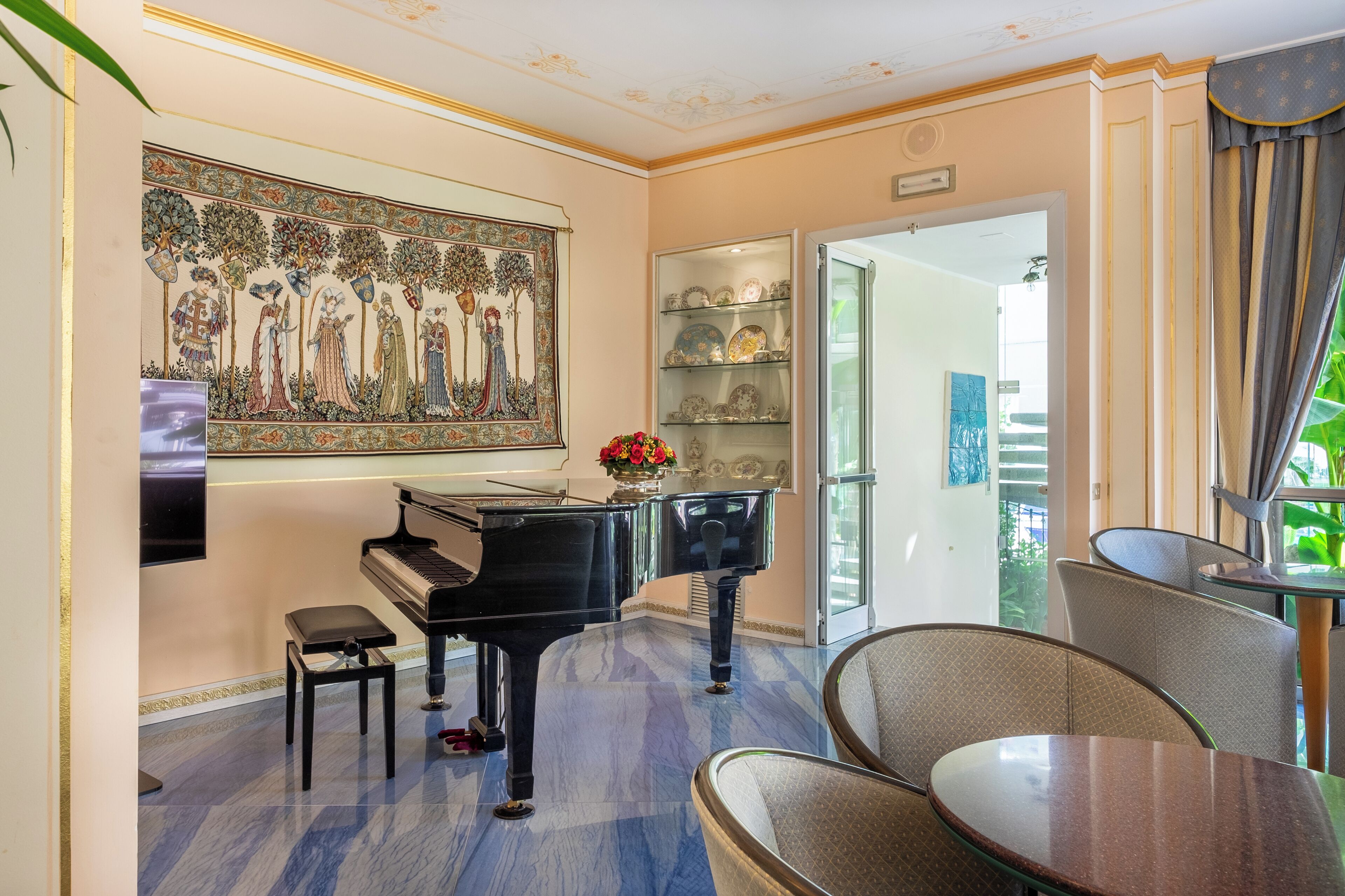 A room with a grand piano, a large decorative painting, a glass display case with china, and a dining area with chairs and a table.