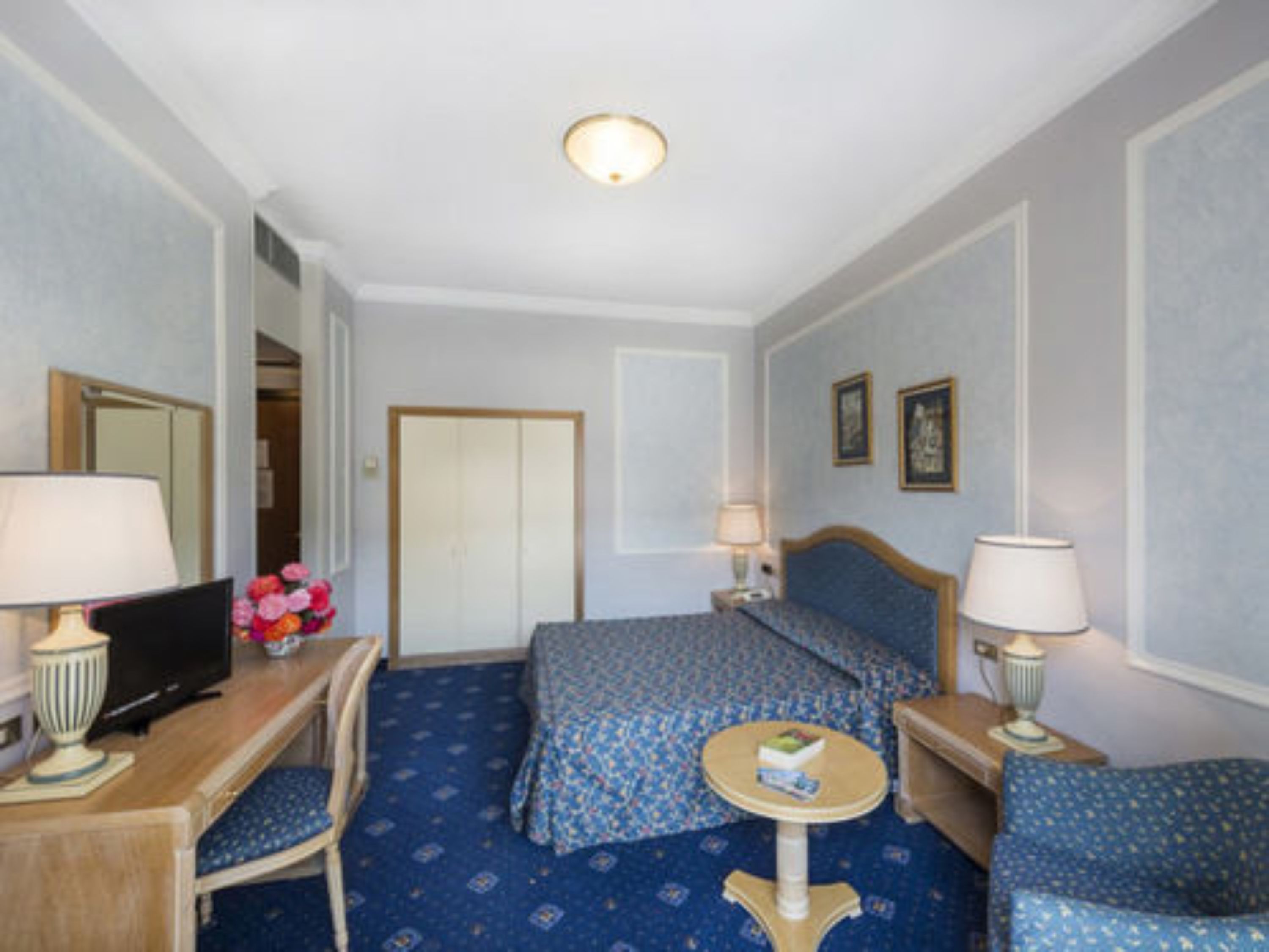 A hotel room with a bed, a desk with a computer, a chair, a small table, and a lamp.