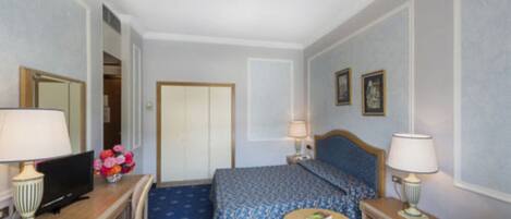 A hotel room with a bed, a desk with a computer, a chair, a small table, and a lamp.