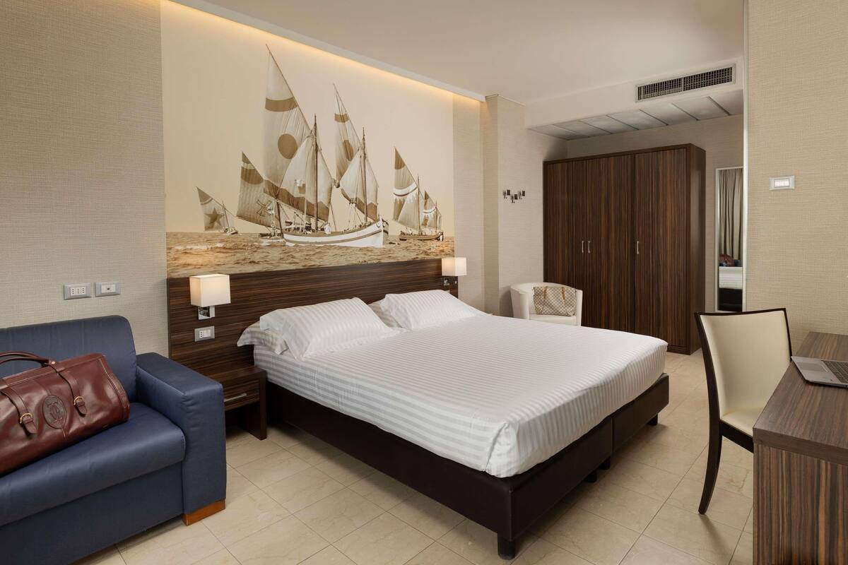 A hotel room with a bed, a blue armchair, a wooden wardrobe, a desk with a laptop, and a wall mural of sailboats.