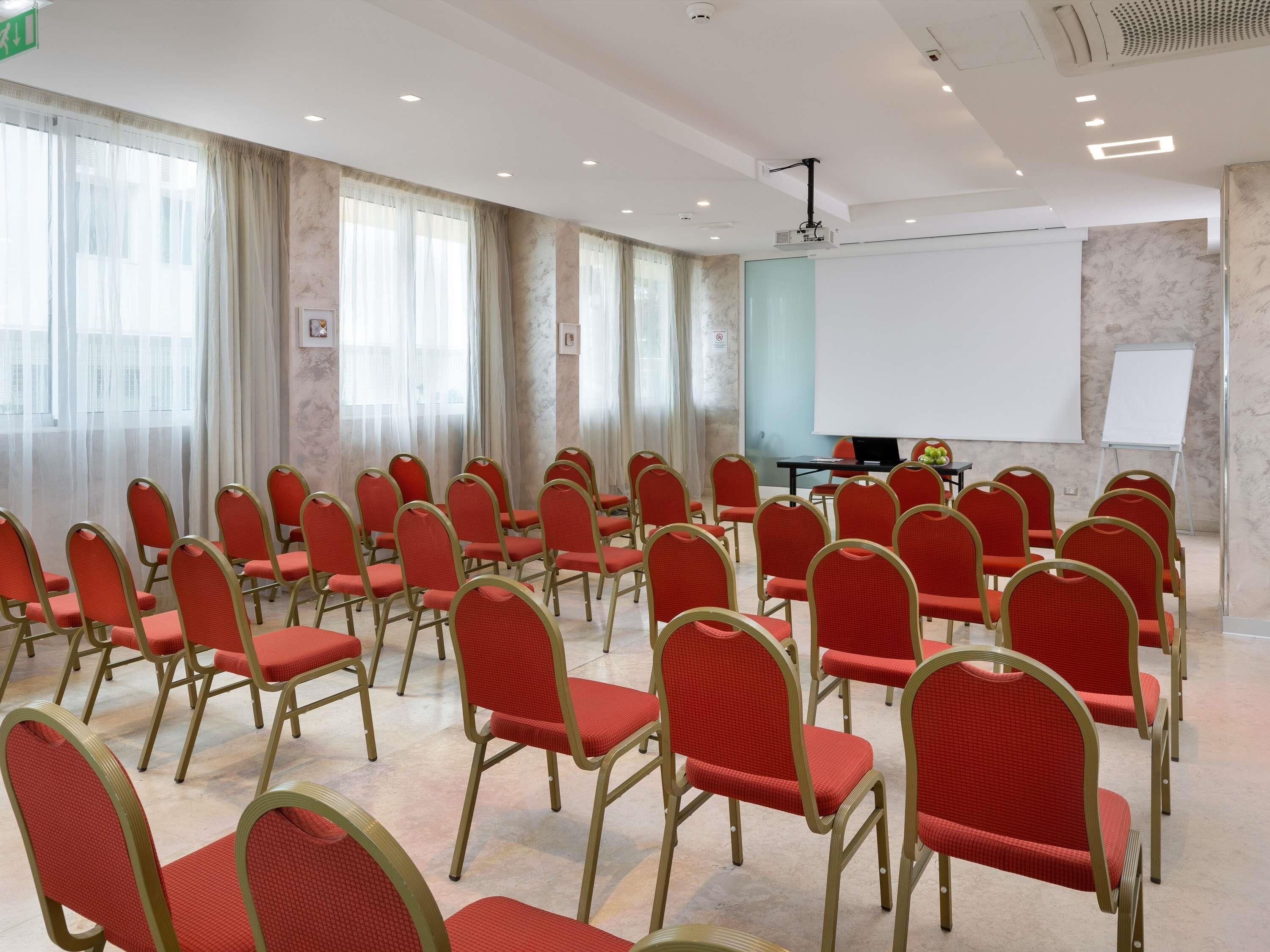 Ein Konferenzraum mit Reihen roter Stühle, einem Projektor, einer Whiteboard und einem großen Fenster mit durchsichtigen Vorhängen.