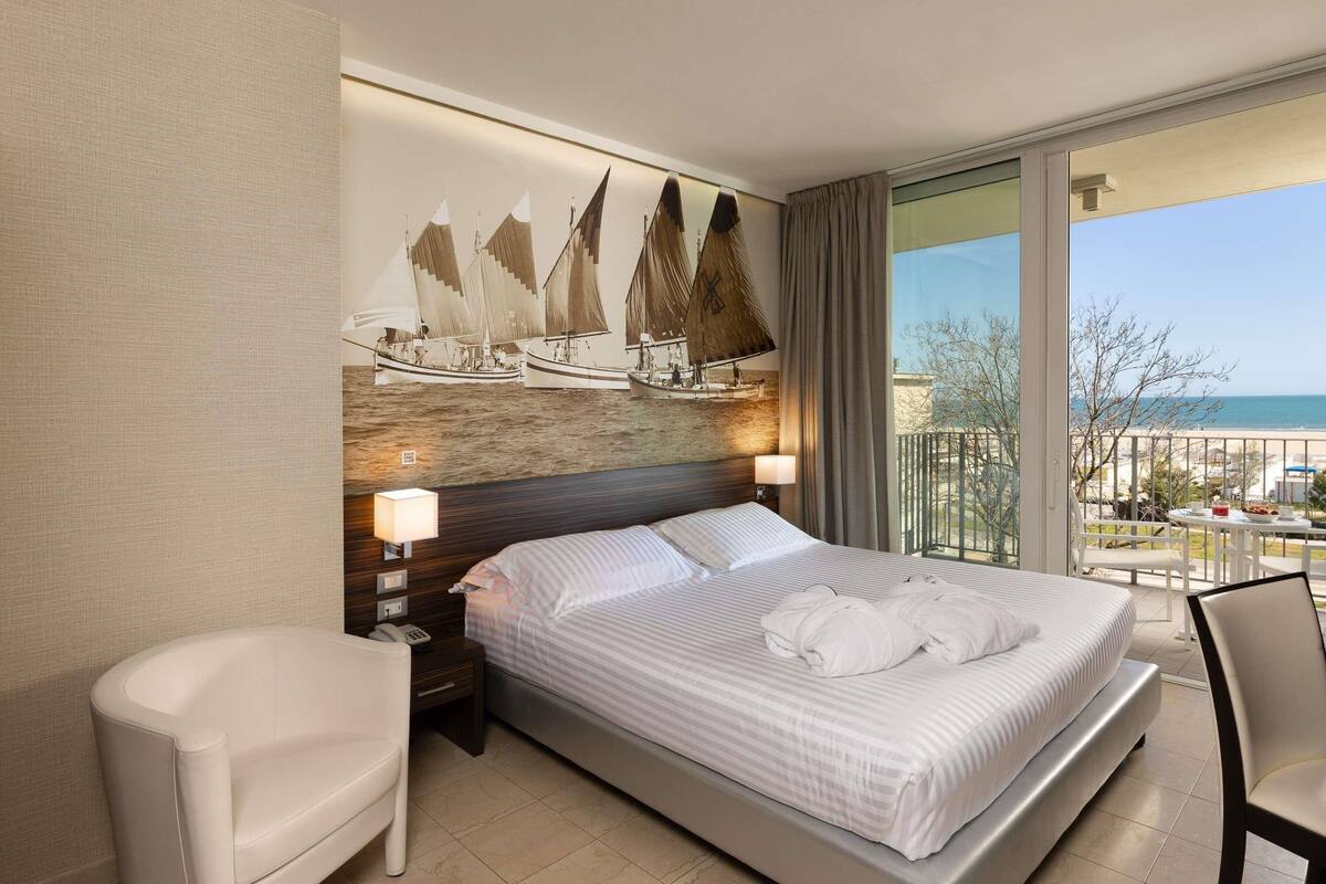 A hotel room with a bed, a chair, a nightstand, a lamp, a painting of sailboats, and a balcony with a view of the sea.