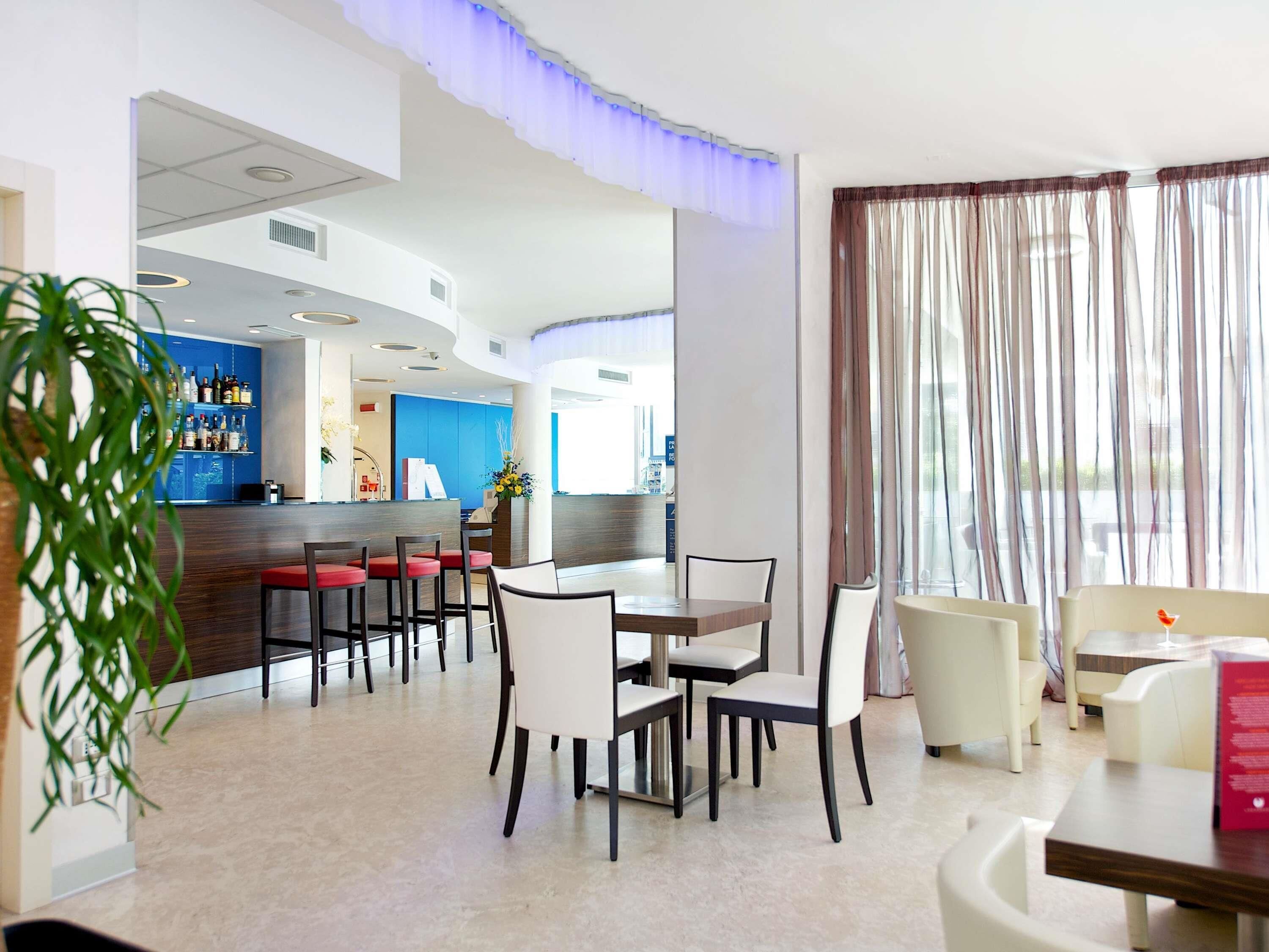 Vestíbulo de hotel con área de bar, disposición de asientos y un gran ventanal con cortinas.