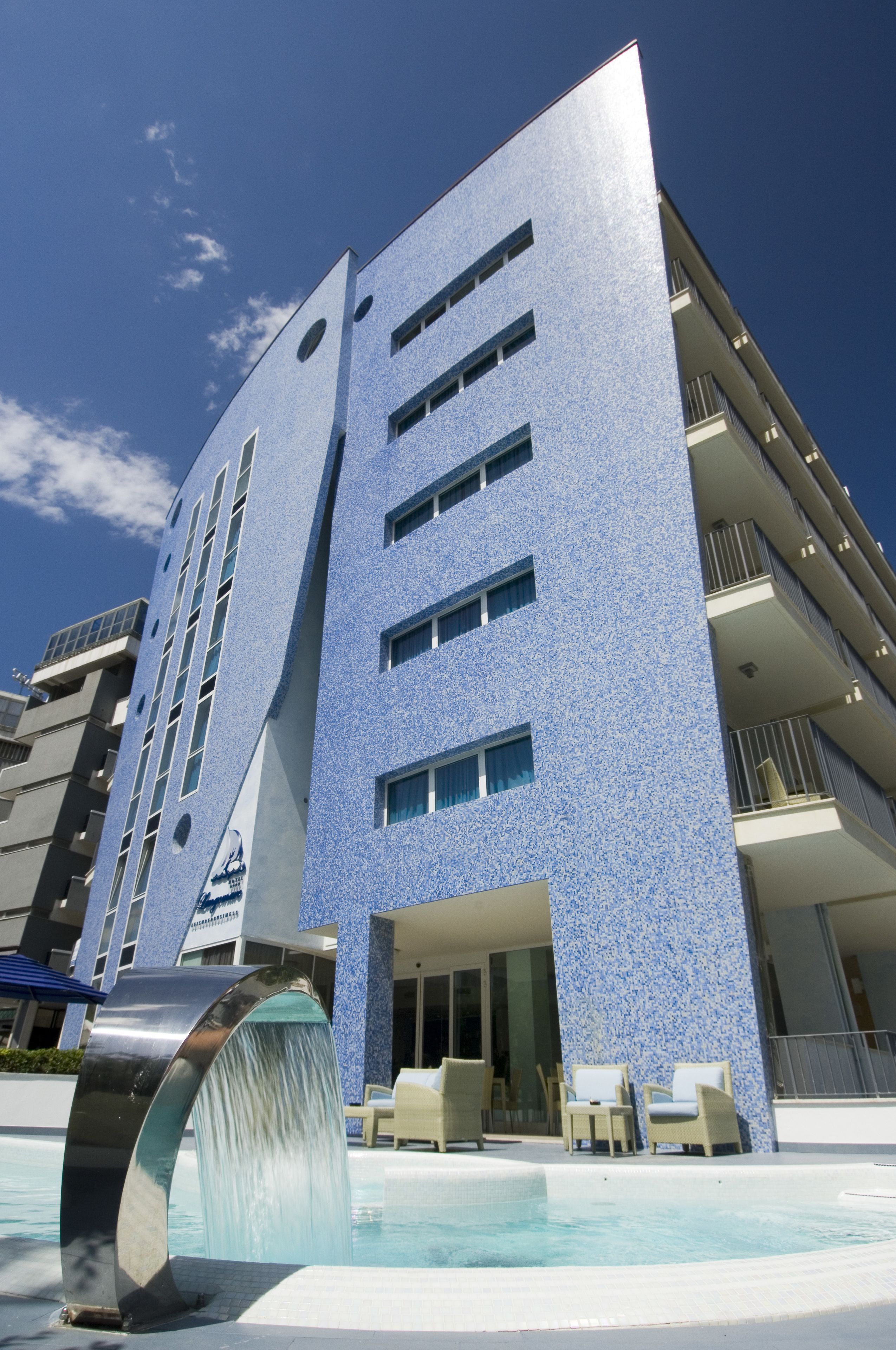 A modern building with a blue textured facade, a pool with a water feature, and outdoor seating.