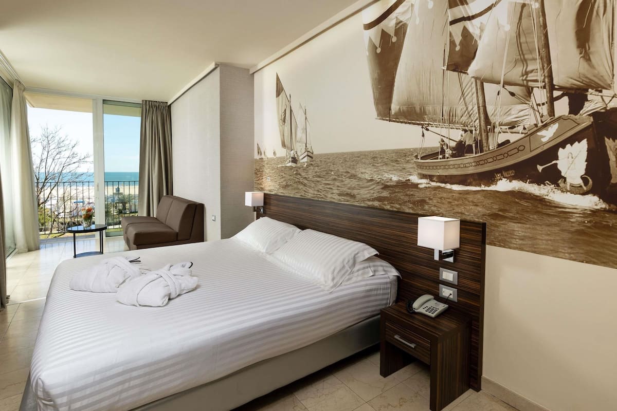 A hotel room with a large bed, a nightstand with a phone, a chair, a table with a vase, and a painting of a sailing ship.