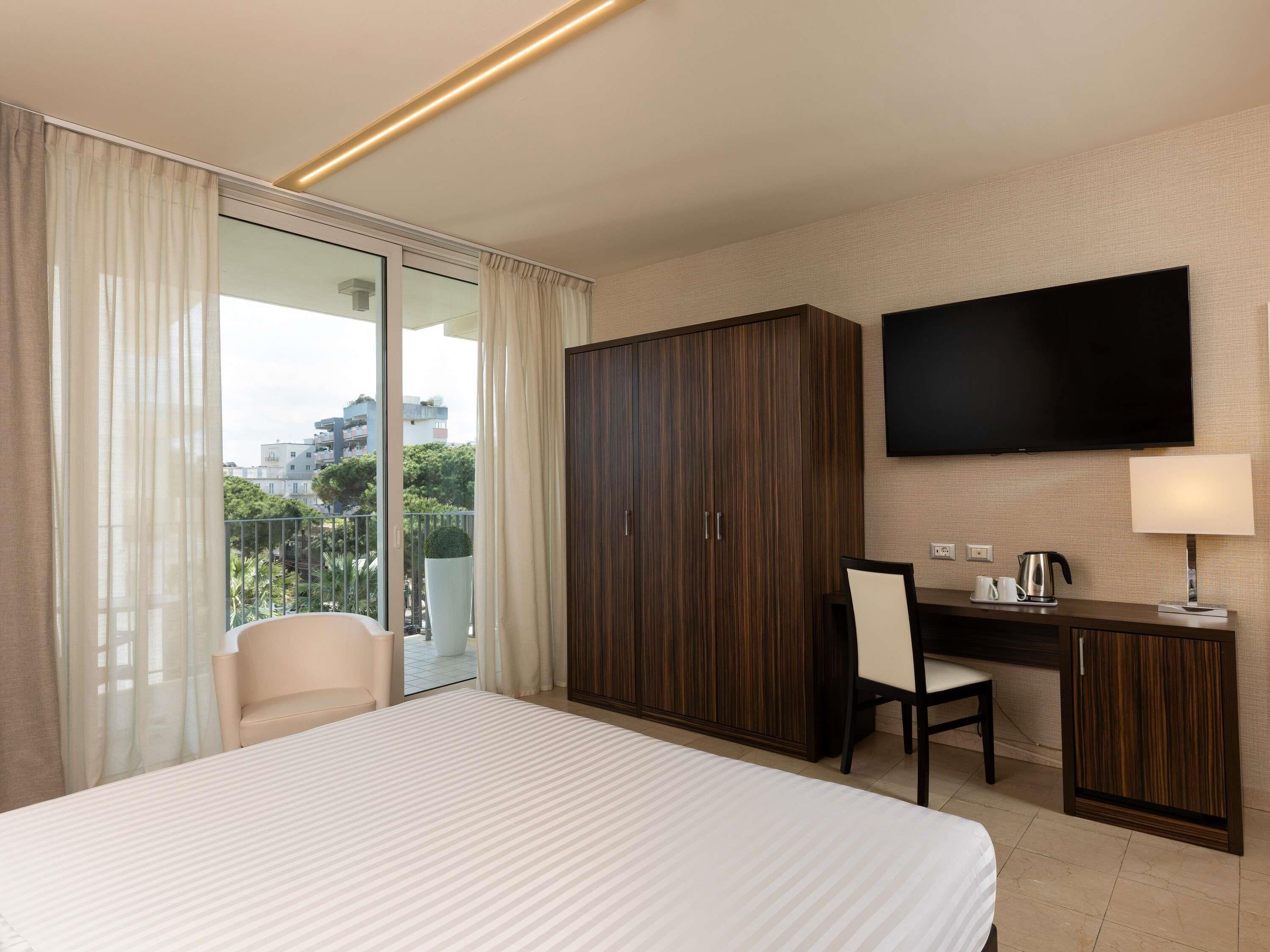 A hotel room with a bed, a desk, a chair, a wardrobe, a TV, and a balcony with curtains.
