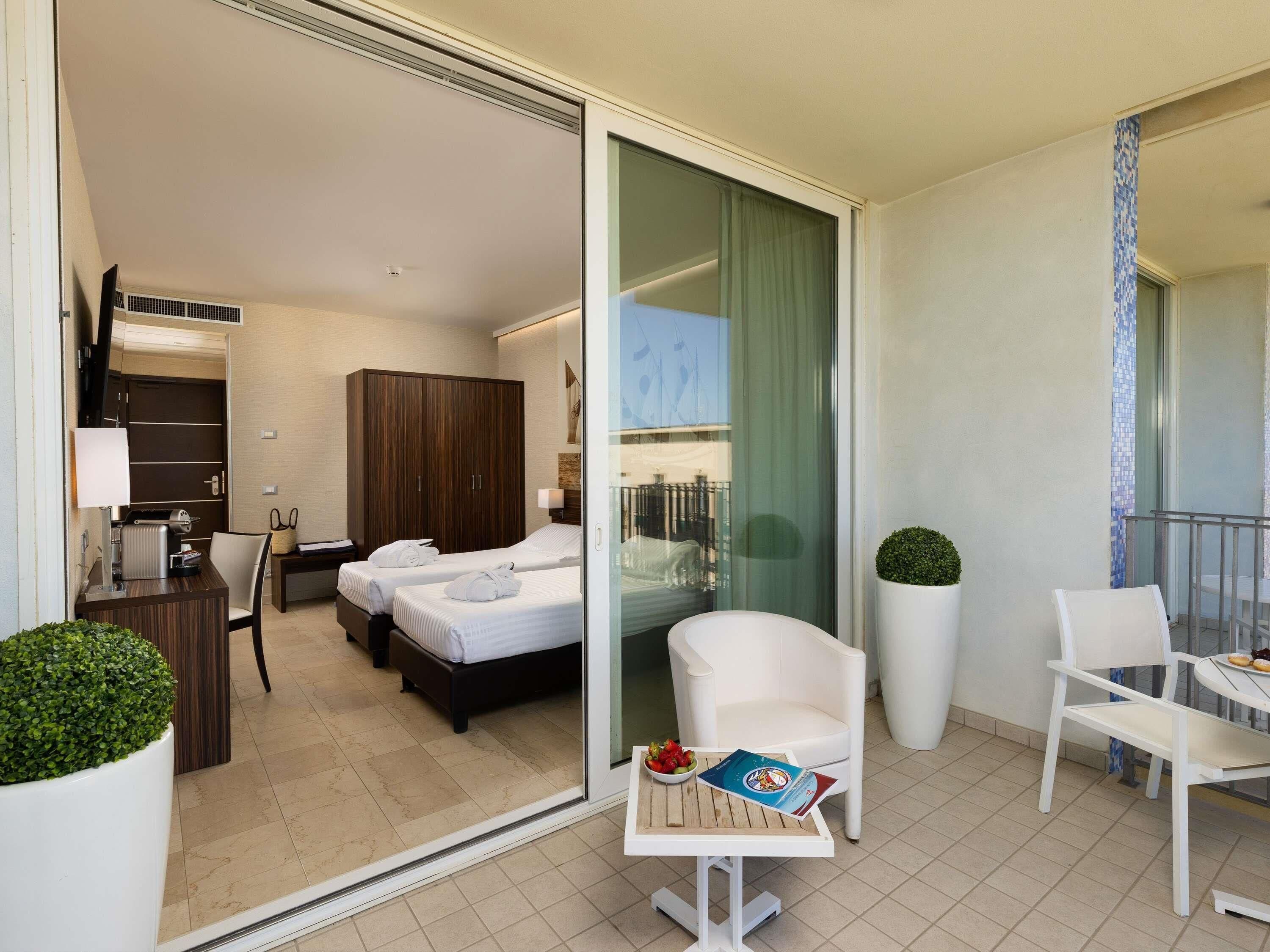 Habitación de hotel con dos camas, un escritorio con lámpara, un armario y un balcón con una silla y mesa.