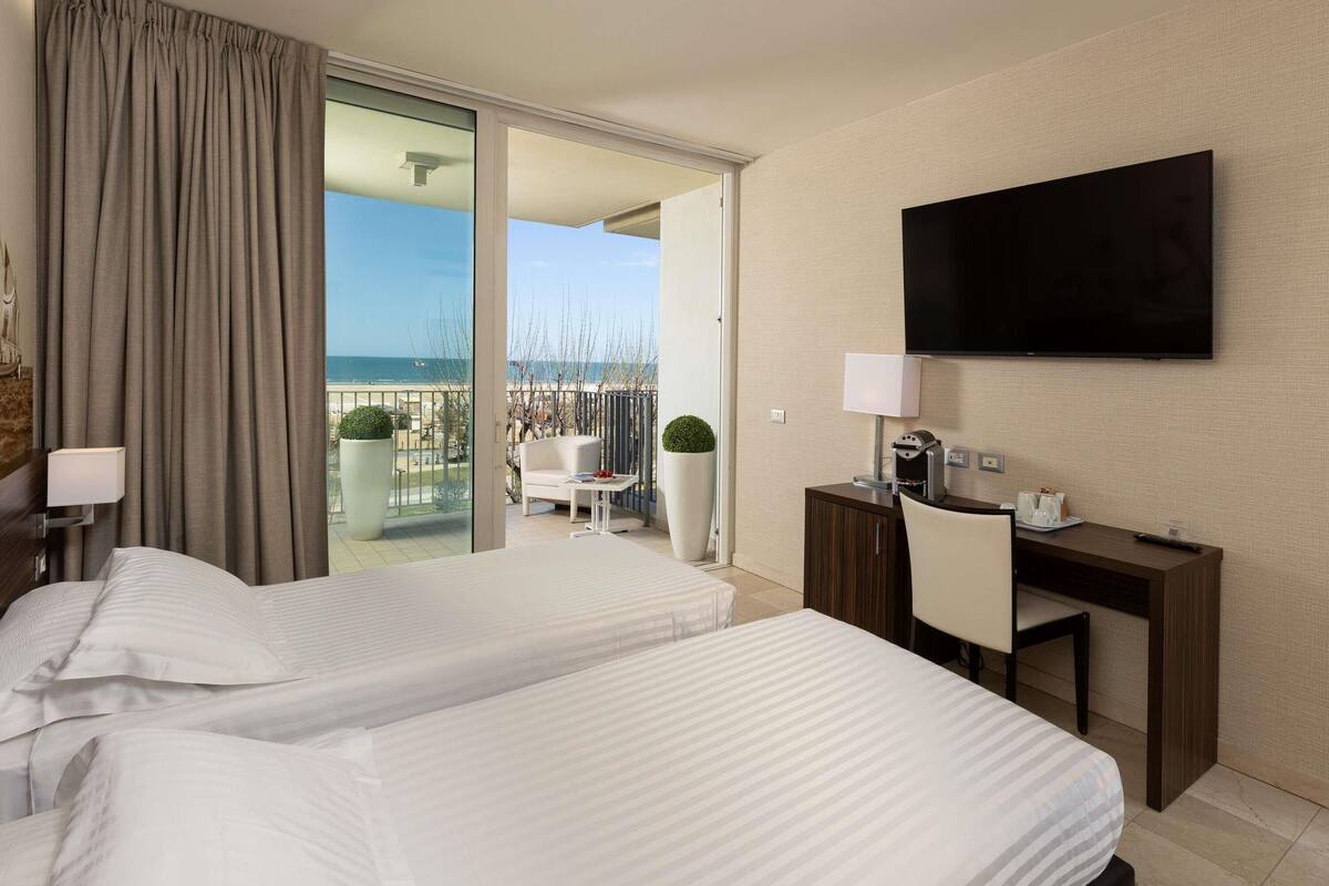 A hotel room with two beds, a desk, a TV, and a balcony with a view of the sea.