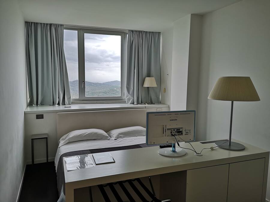 Une chambre d’hôtel avec un lit, un bureau avec un ordinateur, une lampe et une fenêtre avec des rideaux.