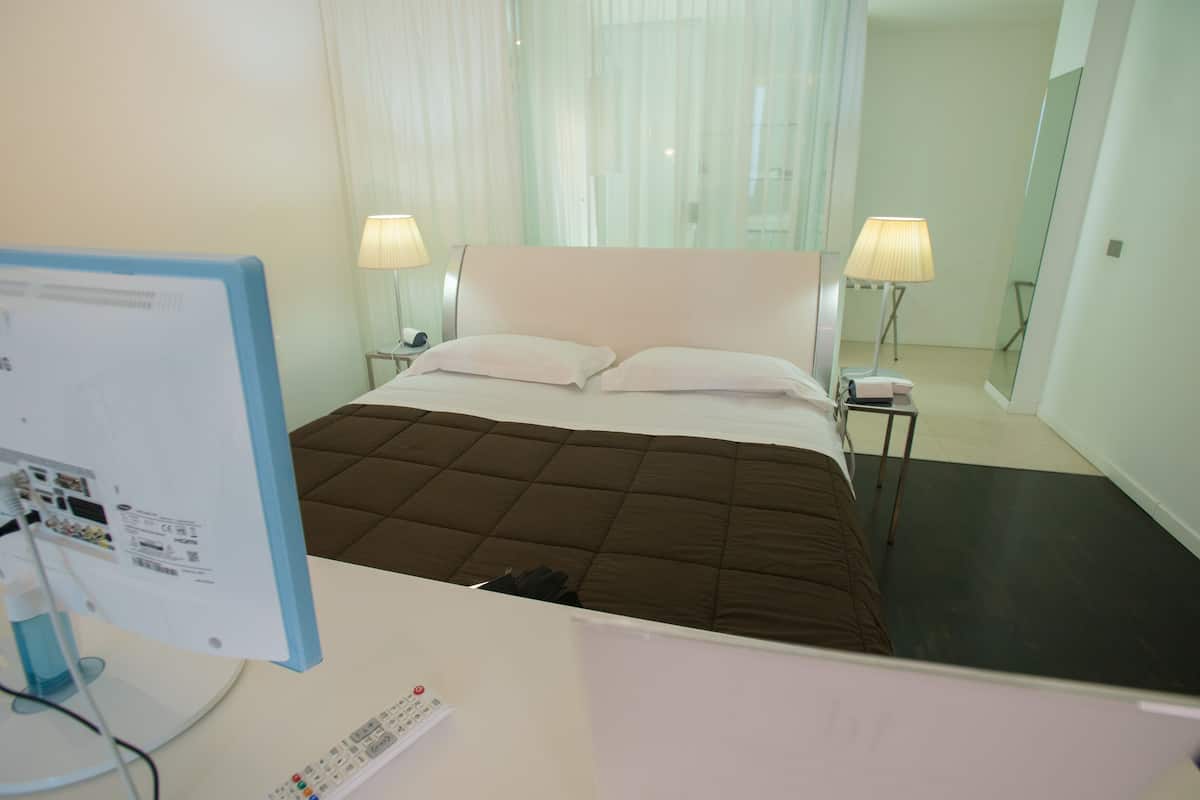 A hotel room with a bed, two bedside lamps, a desk with a computer monitor, and a remote control.