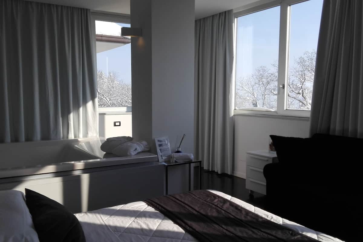 A hotel room with a bed, a bedside table with a lamp, a window with curtains, and a view of trees outside.