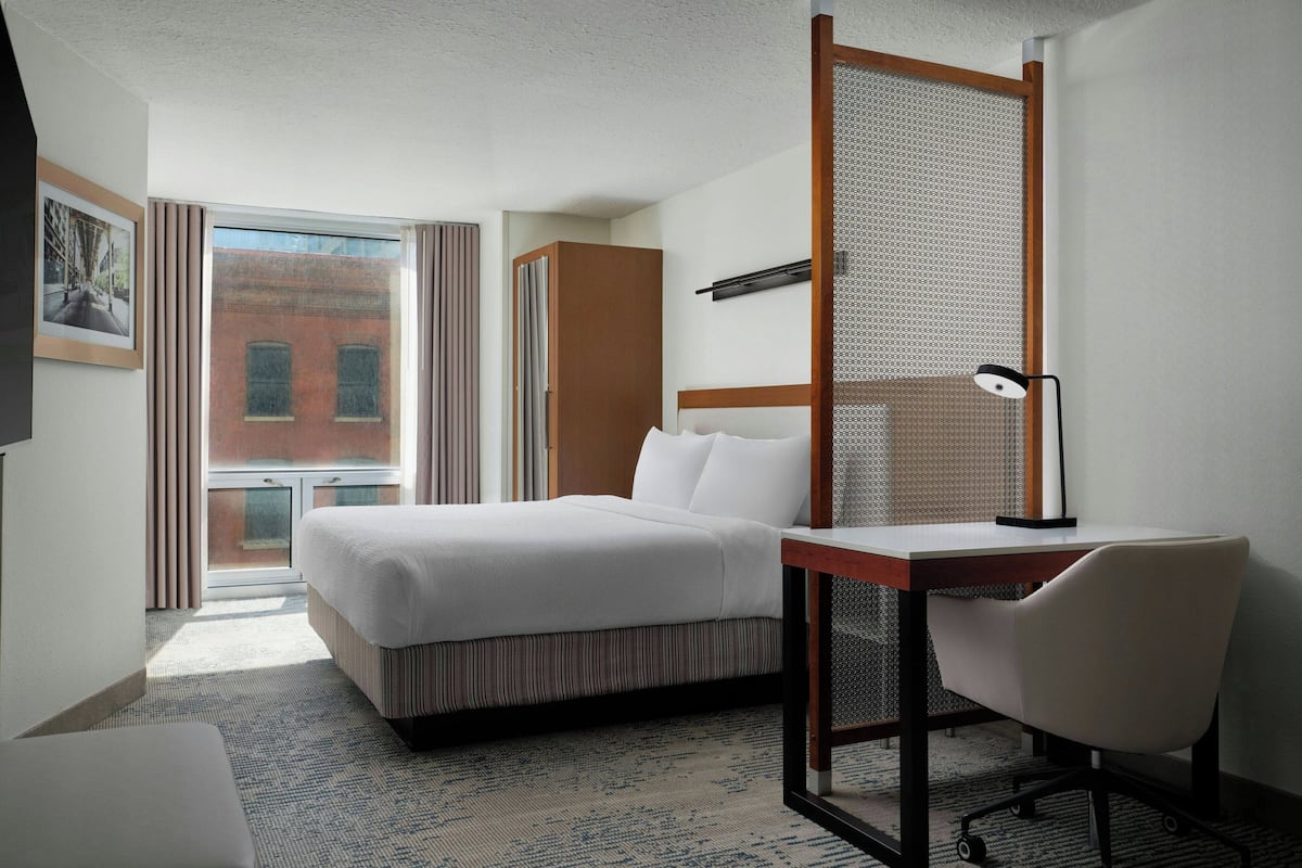 Ein Hotelzimmer mit Bett, Schreibtisch, Stuhl, einem Fenster mit Blick auf ein Gebäude und einer Tür mit Vorhang.