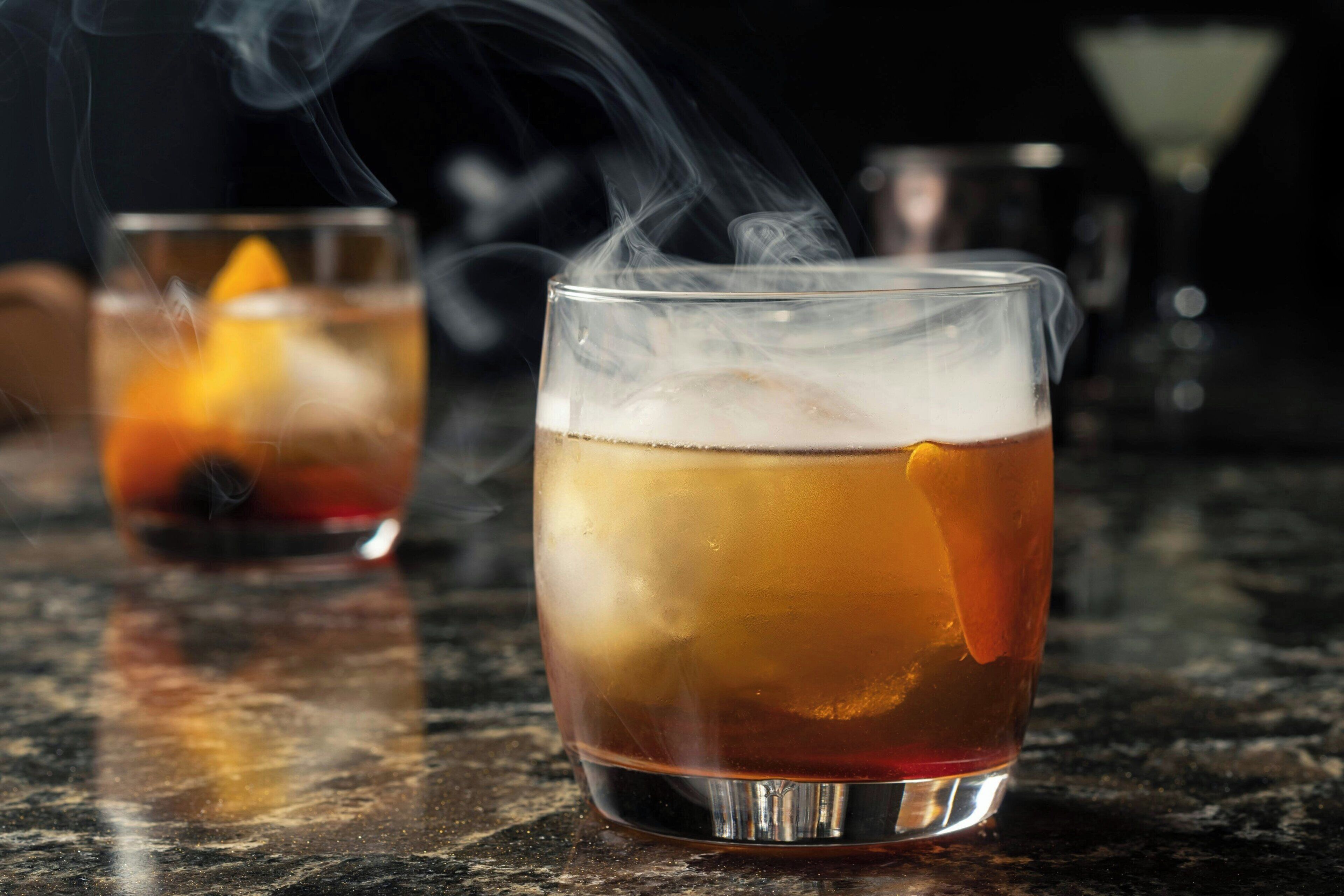 Two glasses of a beverage with a slice of orange, one with smoke rising, on a reflective surface.