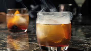 Two glasses of a beverage with a slice of orange, one with smoke rising, on a reflective surface.