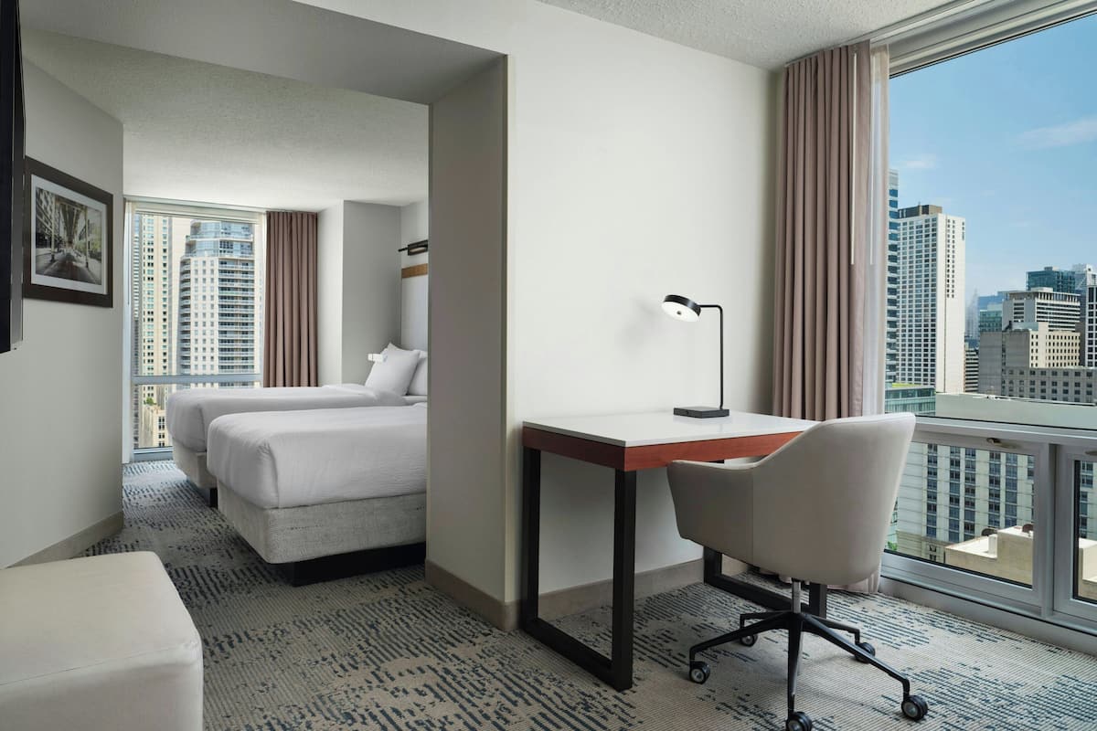 Ein Hotelzimmer mit Bett, Schreibtisch, Stuhl und einem großen Fenster mit Blick auf die Stadt.