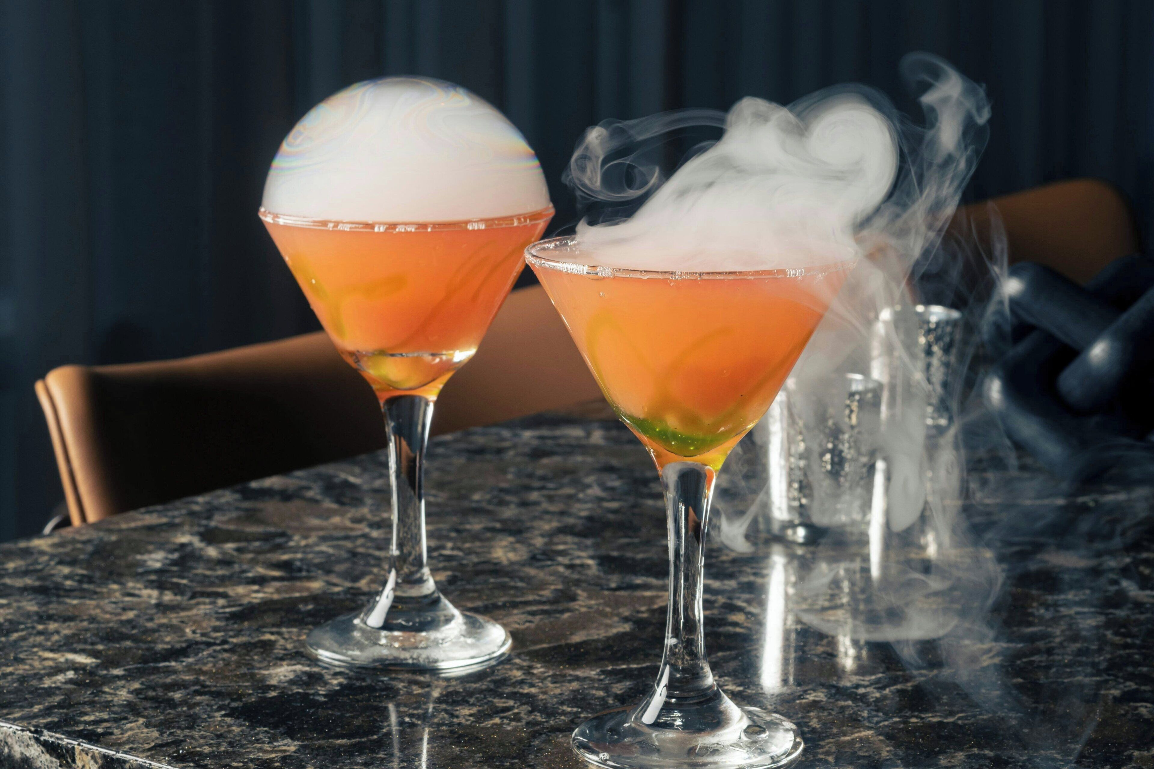 Two cocktails on a marble table with a smoke effect, a chair, and a dark background.