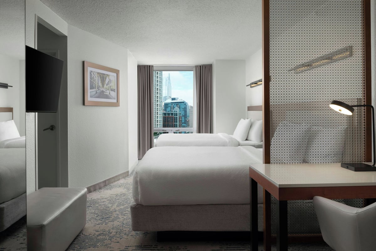 Ein Hotelzimmer mit zwei Betten, einem Schreibtisch, einem Fernseher, Blick auf die Stadt und einem gemusterten Raumteiler.
