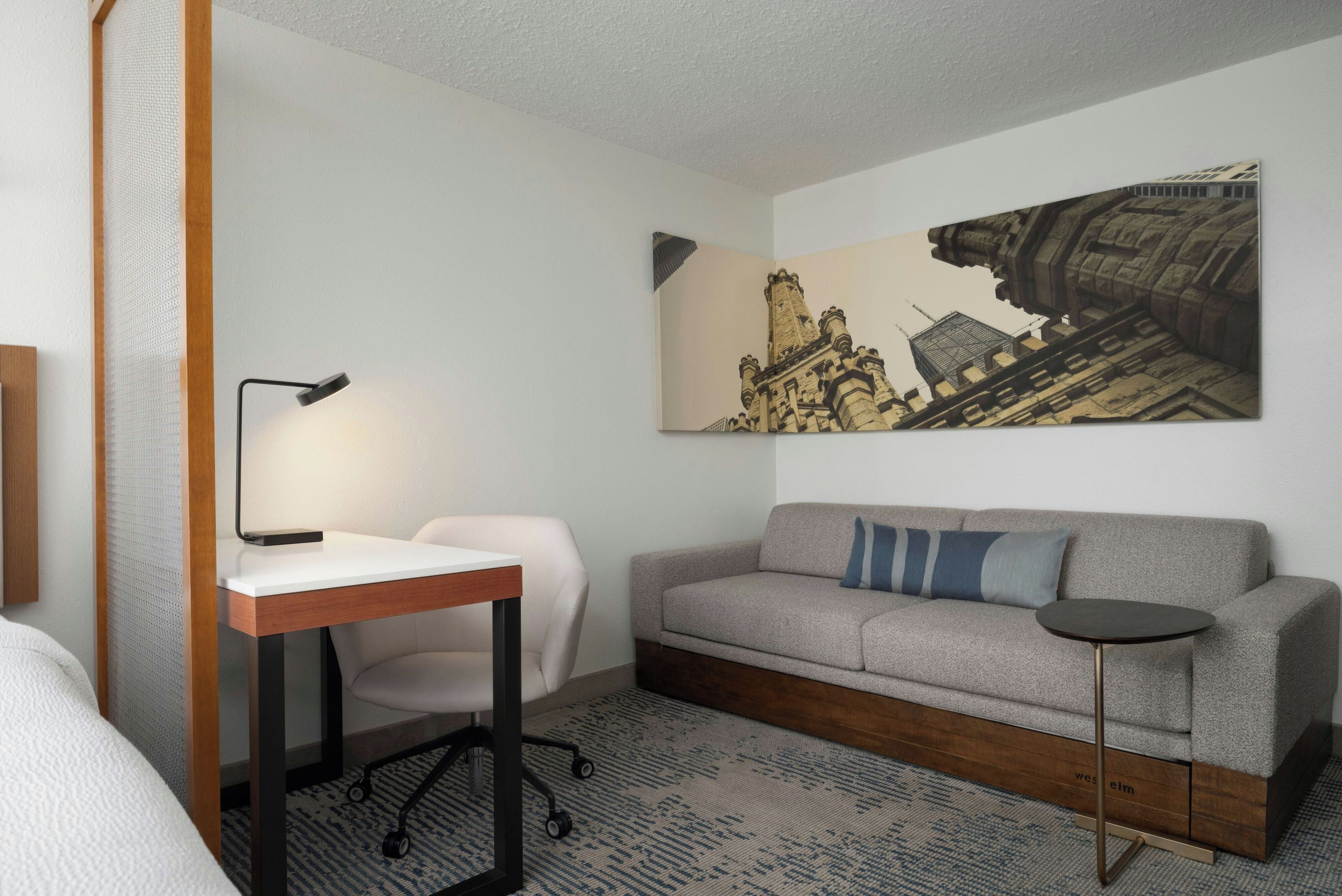 Una camera d'albergo con una scrivania, una sedia, un divano e un grande quadro raffigurante un panorama urbano.