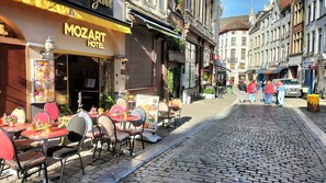 Une rue pavée avec des tables et des chaises installées à l’extérieur du MOZART HOTEL, agrémentée d’une enseigne et de quelques plantes en pot.